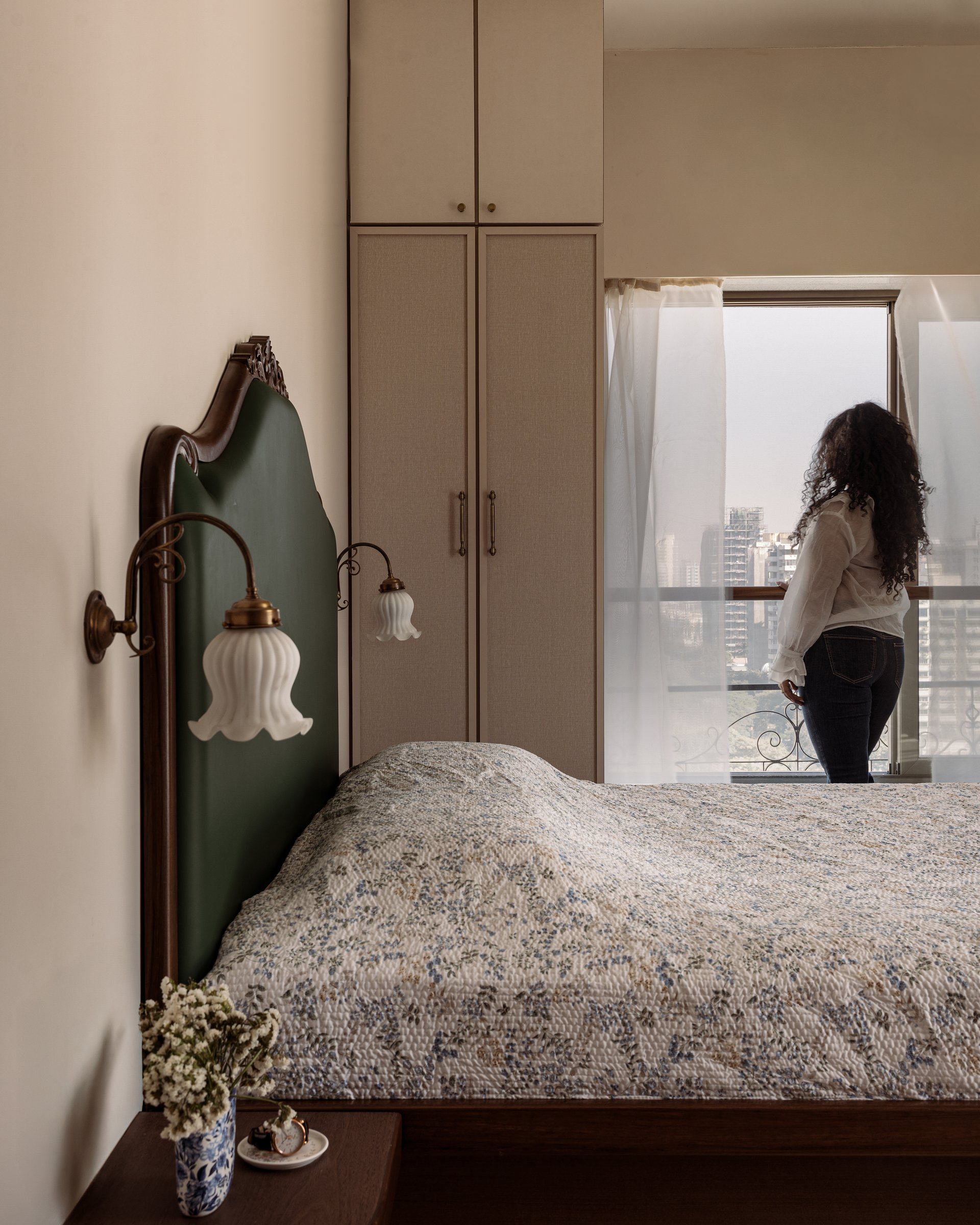 The bedroom, anchored by a forest-green upholstered headboard, brass tulip sconces, and a fabric-fronted wardrobe in oat tones