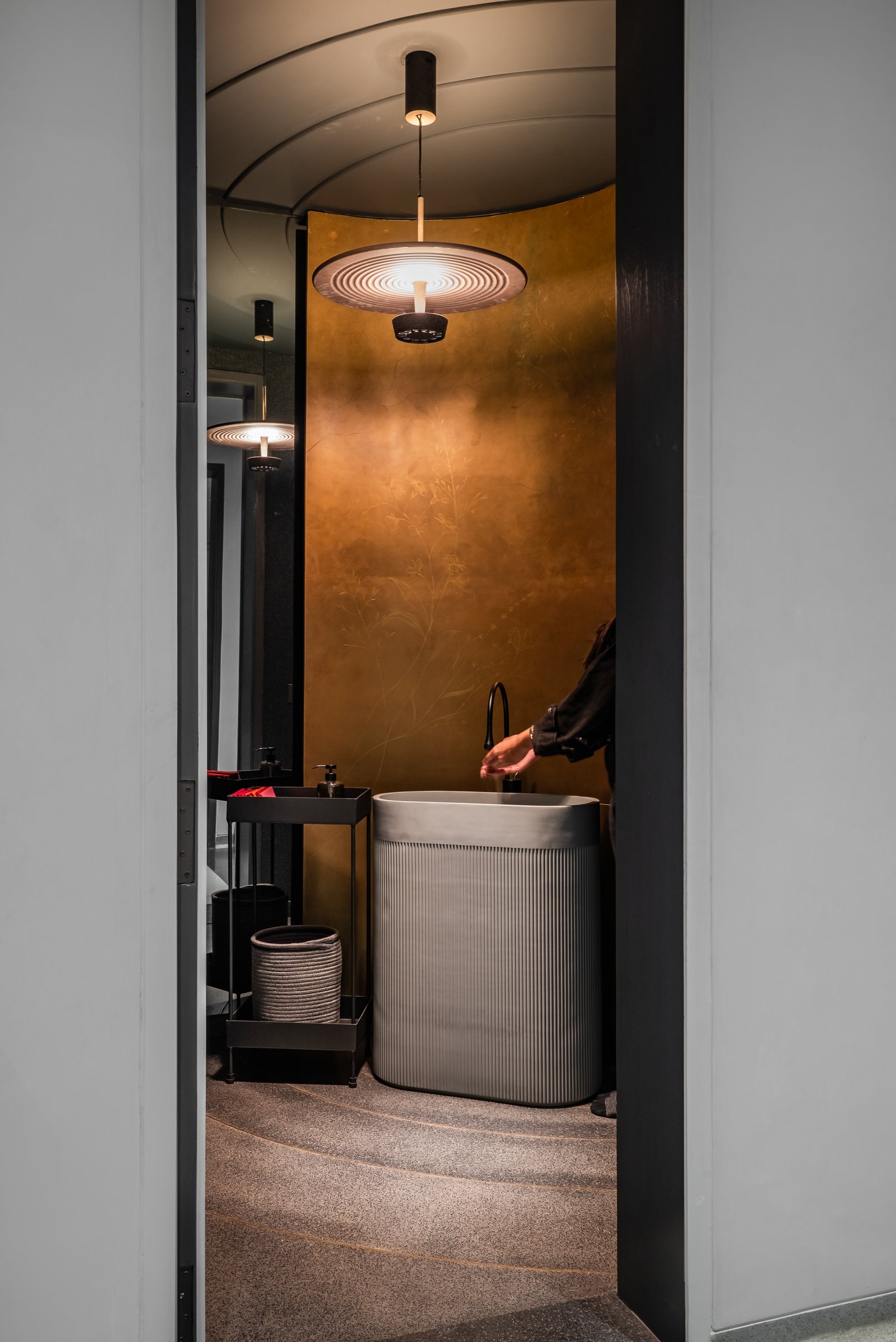A powder room wrapped in a curved metallic-bronze backdrop, lit by a single ringed pendant