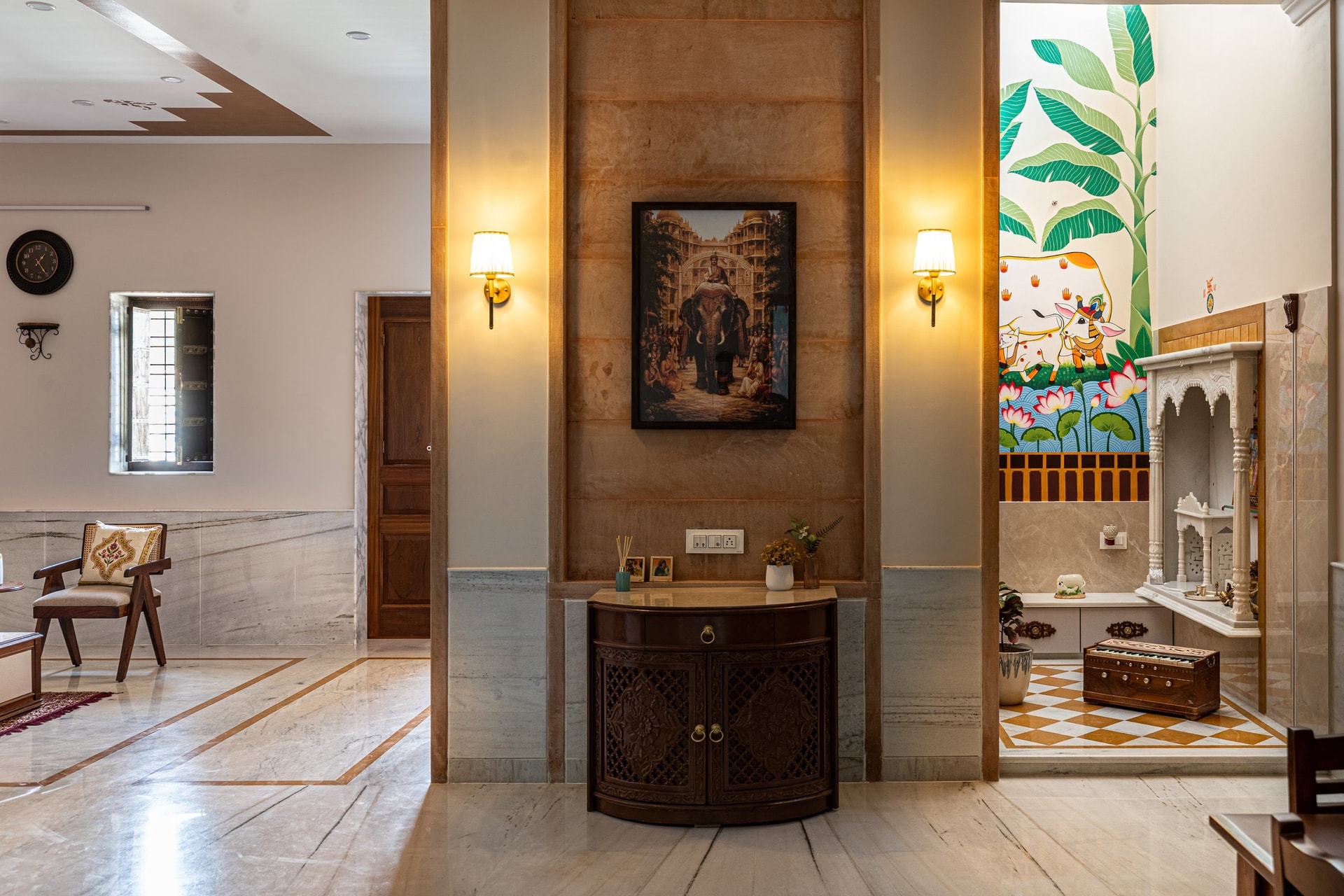 The transition zone between living and pooja room, sandstone pier and dark-wood console on one side, Pichwai mural and marble shrine on the other