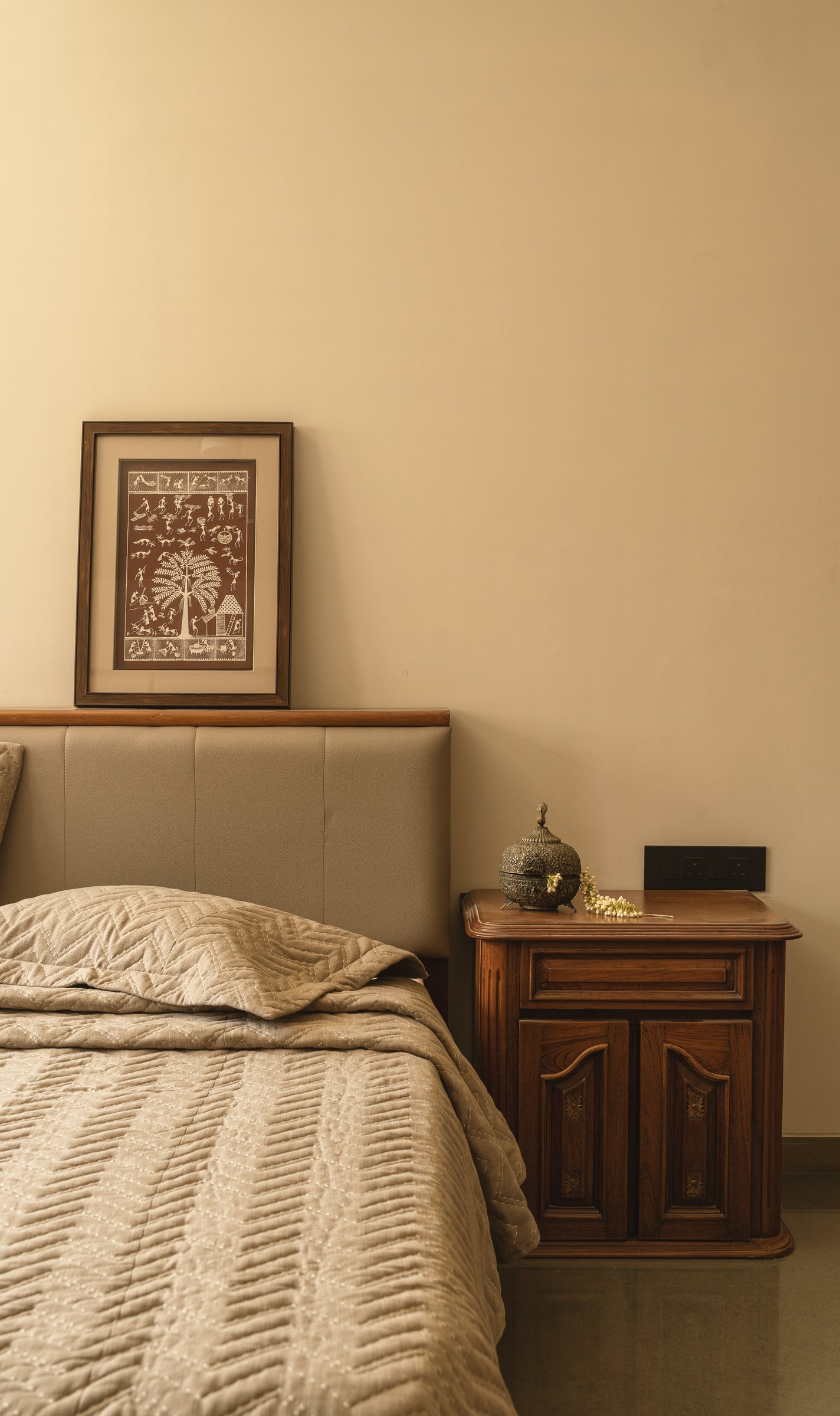A framed Warli painting and a carved bedside cabinet treat regional folk art and inherited furniture as primary, not accessory