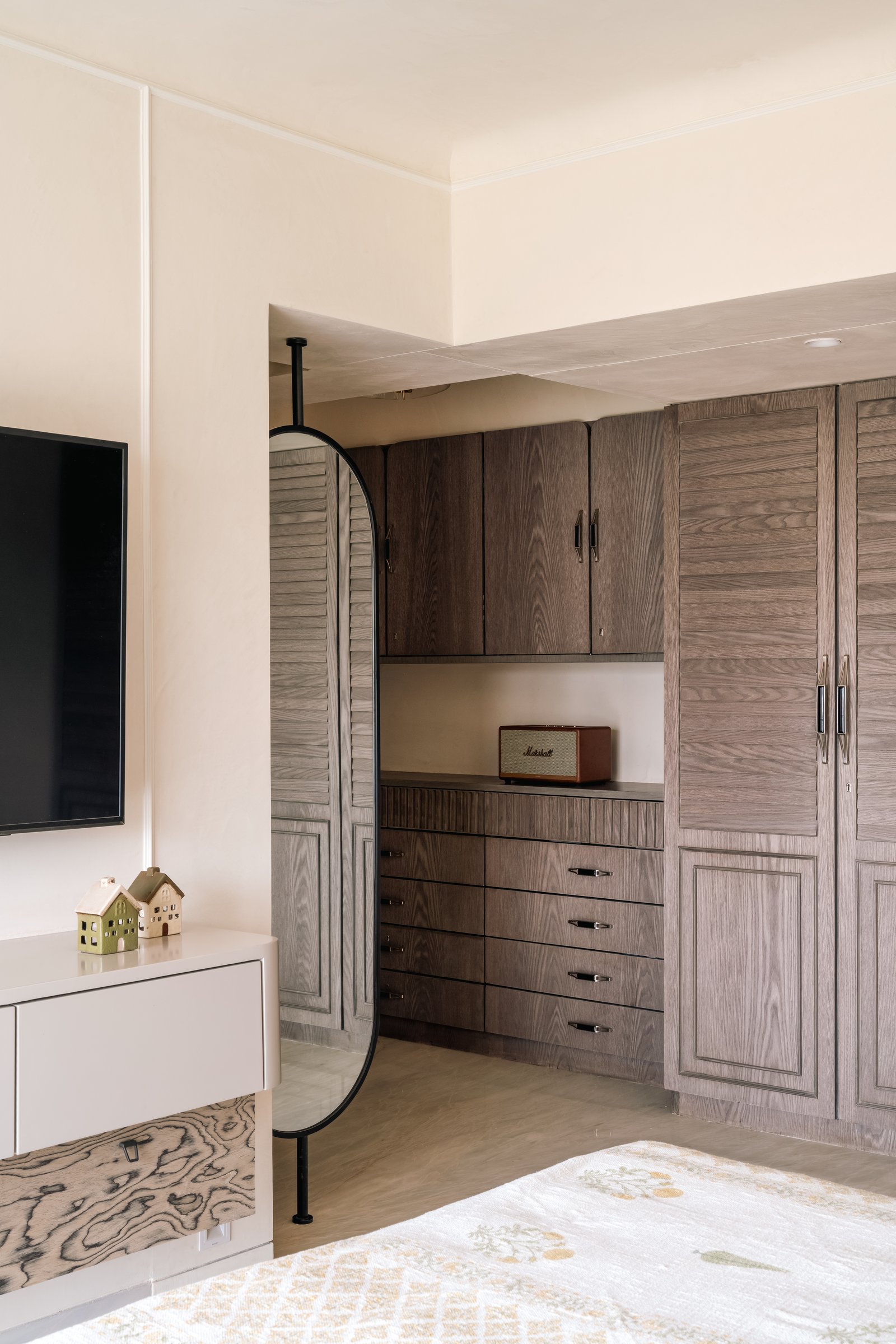 The master's storage wall: louvred and panelled fumed-wood doors, fluted drawer fronts, and an oval freestanding mirror on a slim black armature