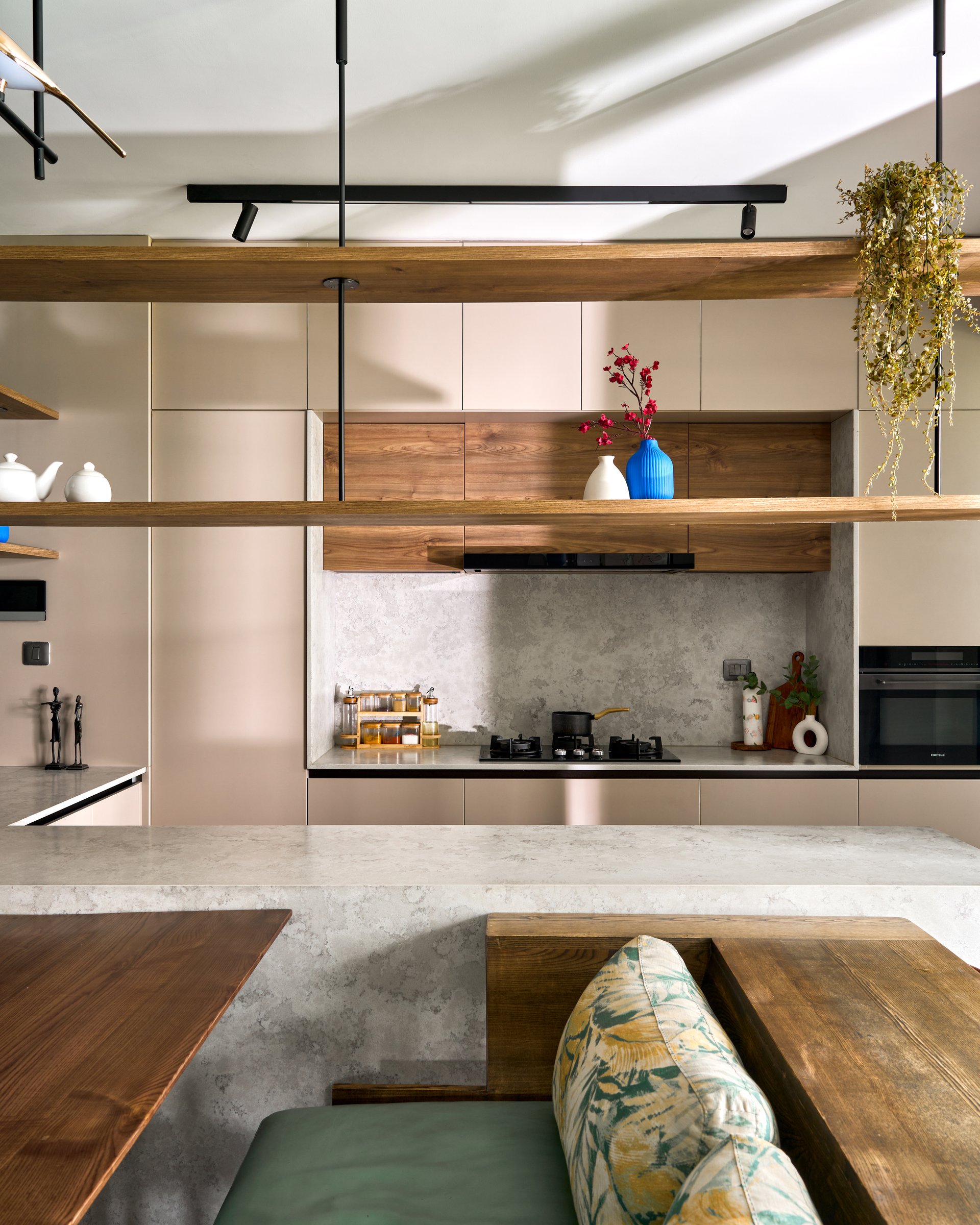 The kitchen seen from the banquette, layered in blush cabinetry, floating wood shelves, and a stone backsplash