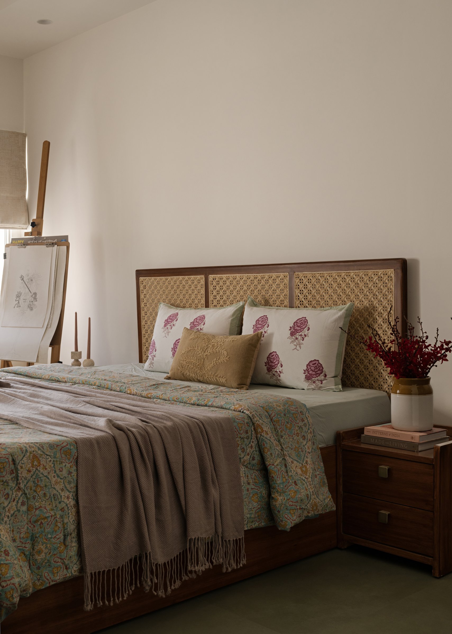 A wide cane-webbed headboard set against bare plaster, a painter's easel standing in the corner