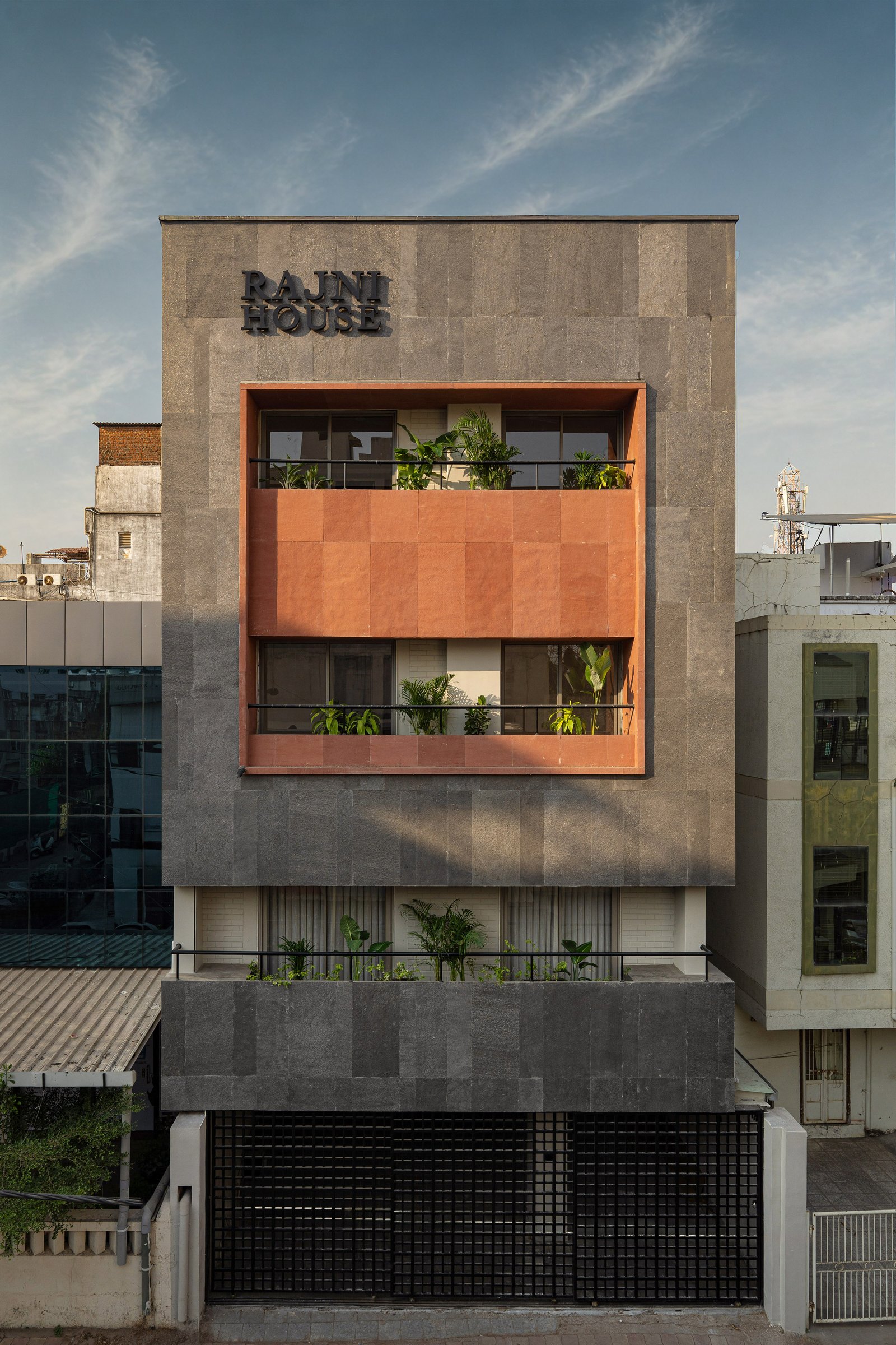 The renewed facade, where a grey stone field frames a recessed terracotta-toned volume planted with tropical foliage