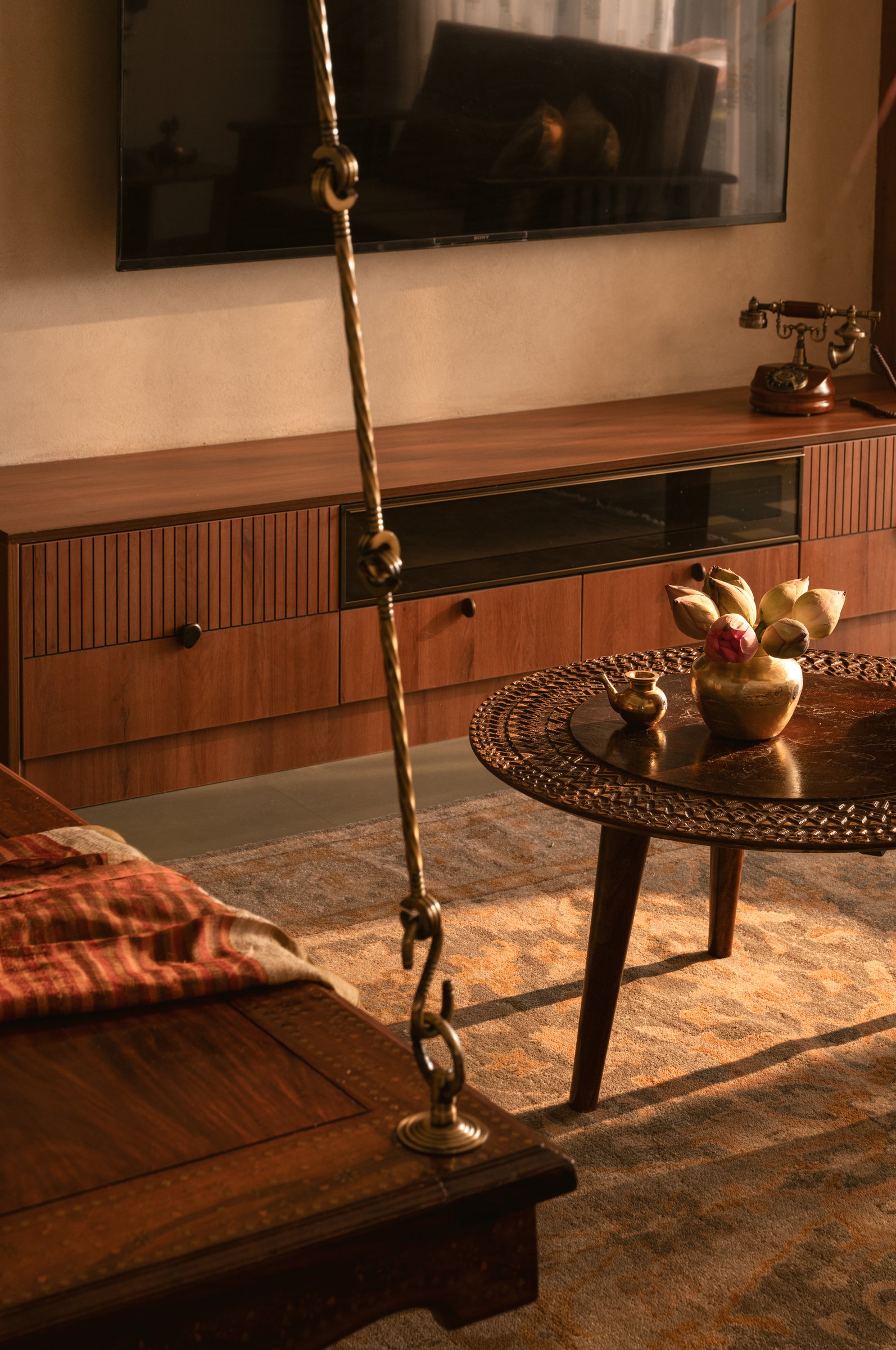 The media console in reeded wood, the carved tripod table beside it carrying a single brass vessel of lotus buds