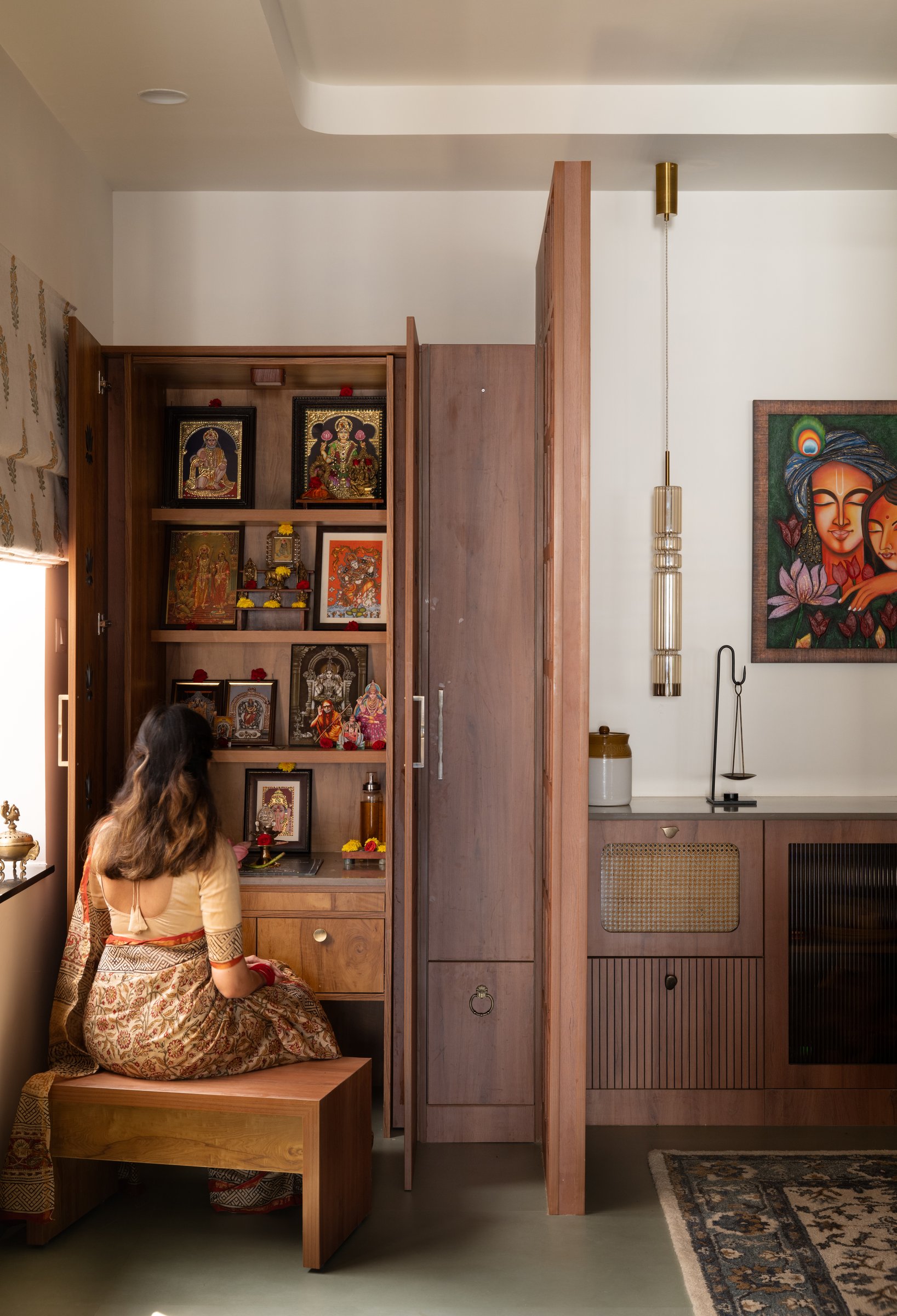 The pooja cabinet open, with stepped shelves of Tanjore paintings and brass idols framed by a small bench for darshan