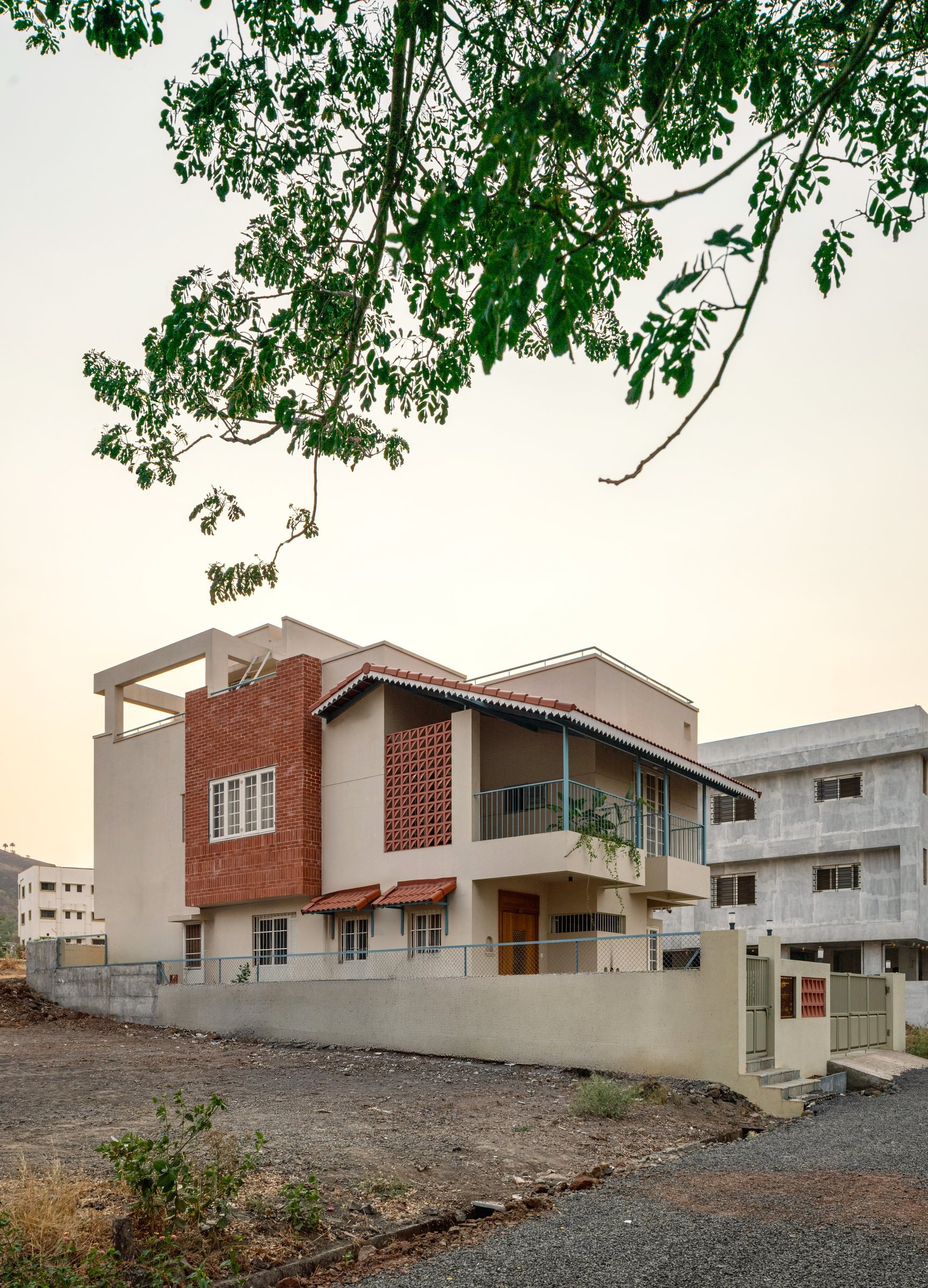 The street view at dusk: a cream stucco mass broken by a brick volume and a perforated terracotta jaali, with the Mangalore-tile awning establishing the regional register