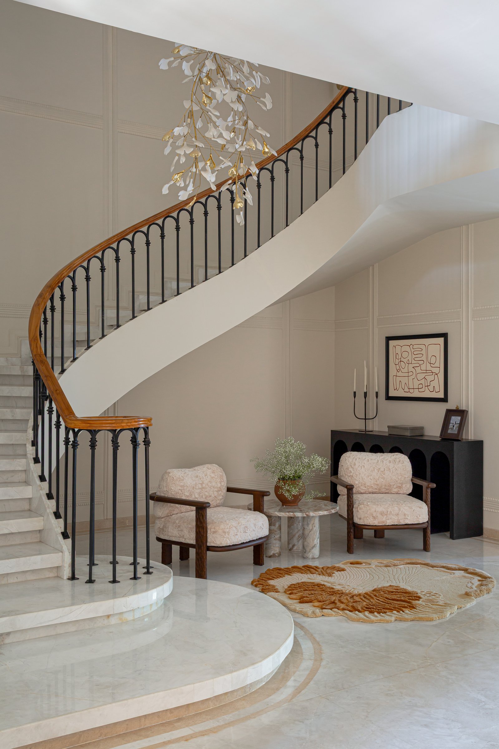 The entry and staircase: a sculptural sweep of stone held by a wrought-iron balustrade, beneath a chandelier of porcelain ginkgo leaves and brass branches