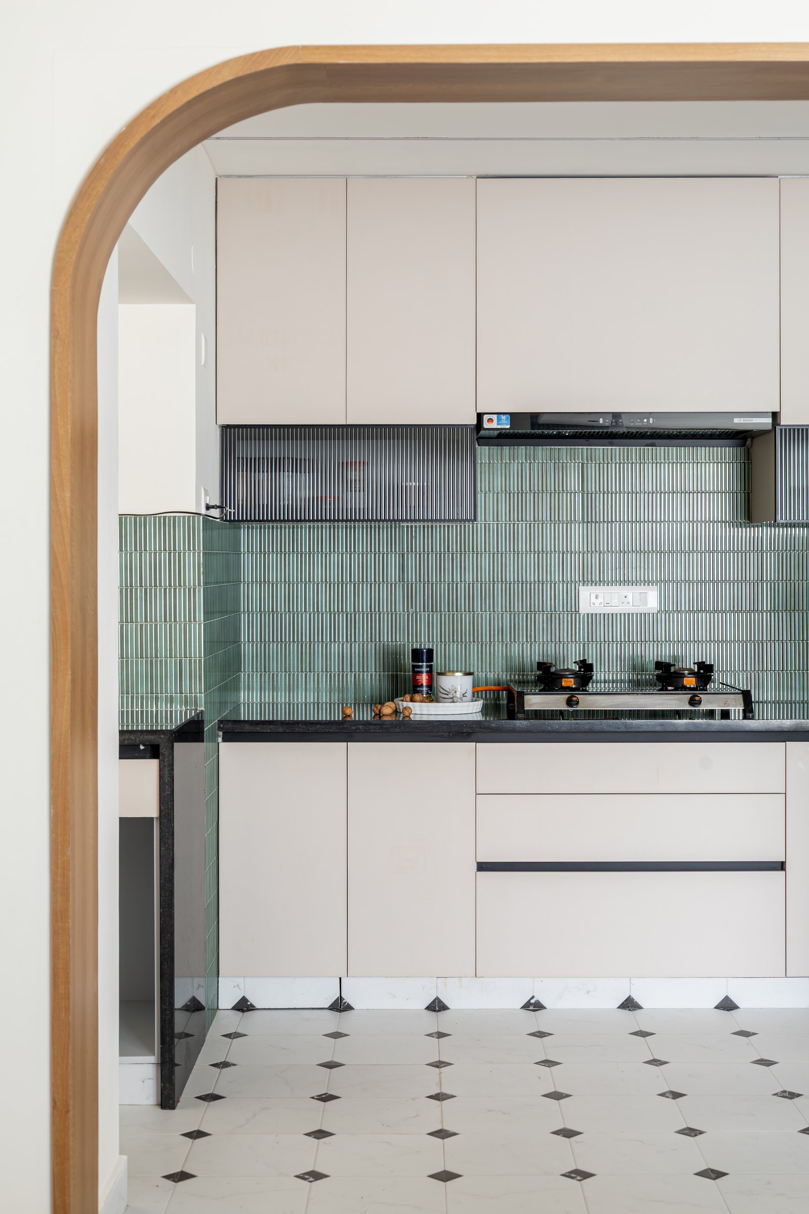 The kitchen seen through a curved oak threshold: sage-green kitkat tile against blush cabinetry