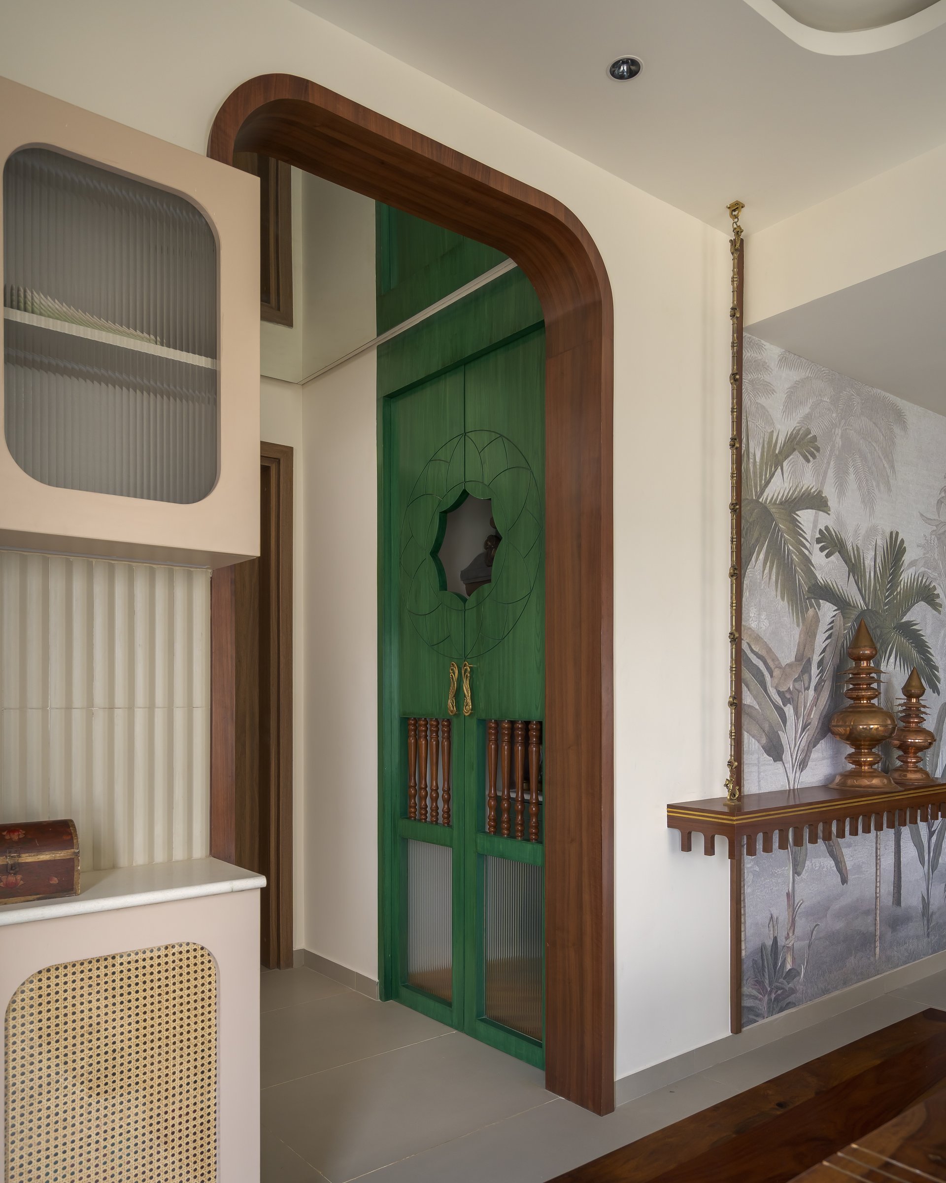 The transition threshold, where an arched wooden portal frames a green pooja door beside a mural-and-console vignette