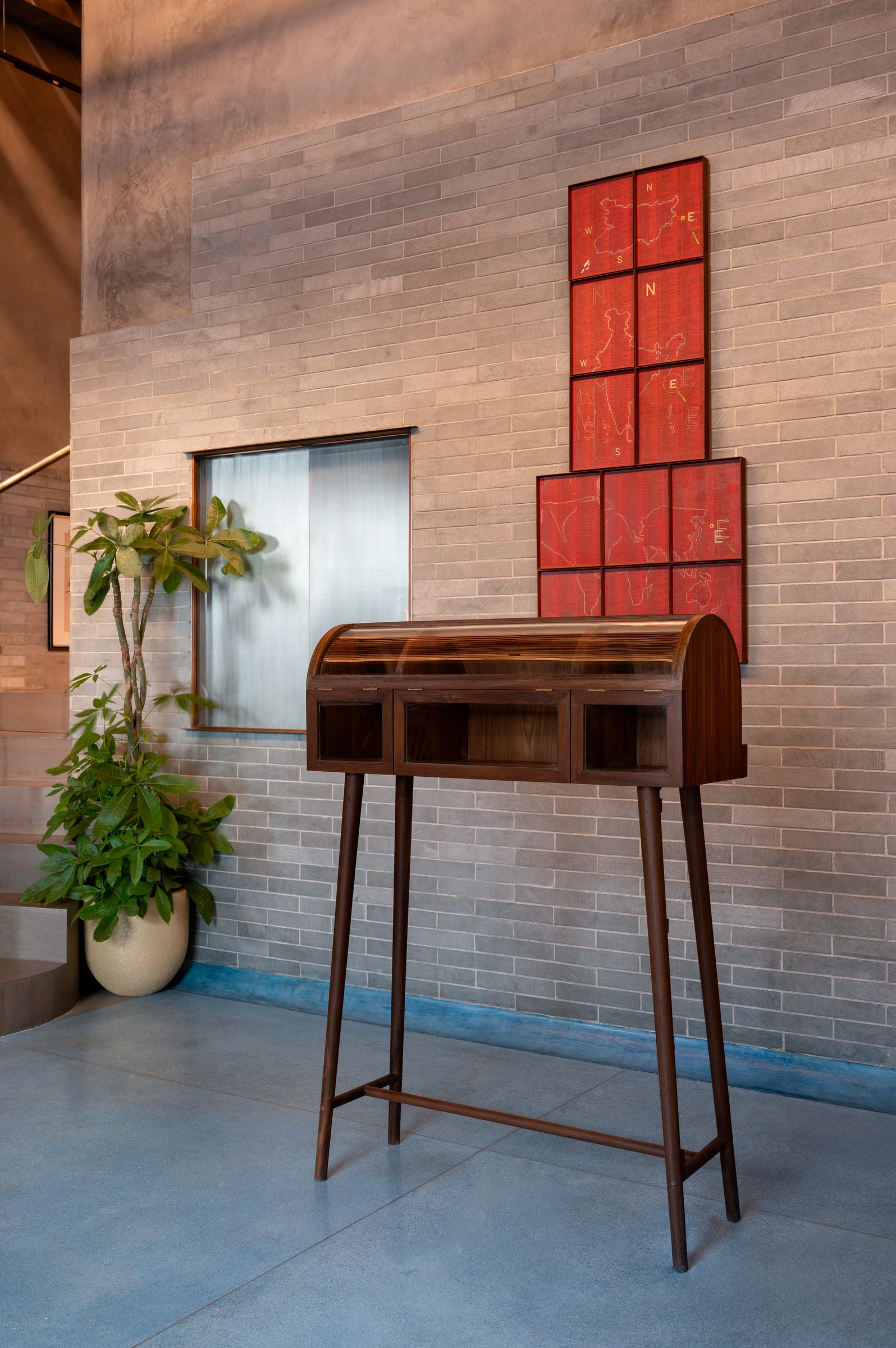 A slim wooden bureau on tapered legs, crowned by a grid of red-toned framed cartographic works