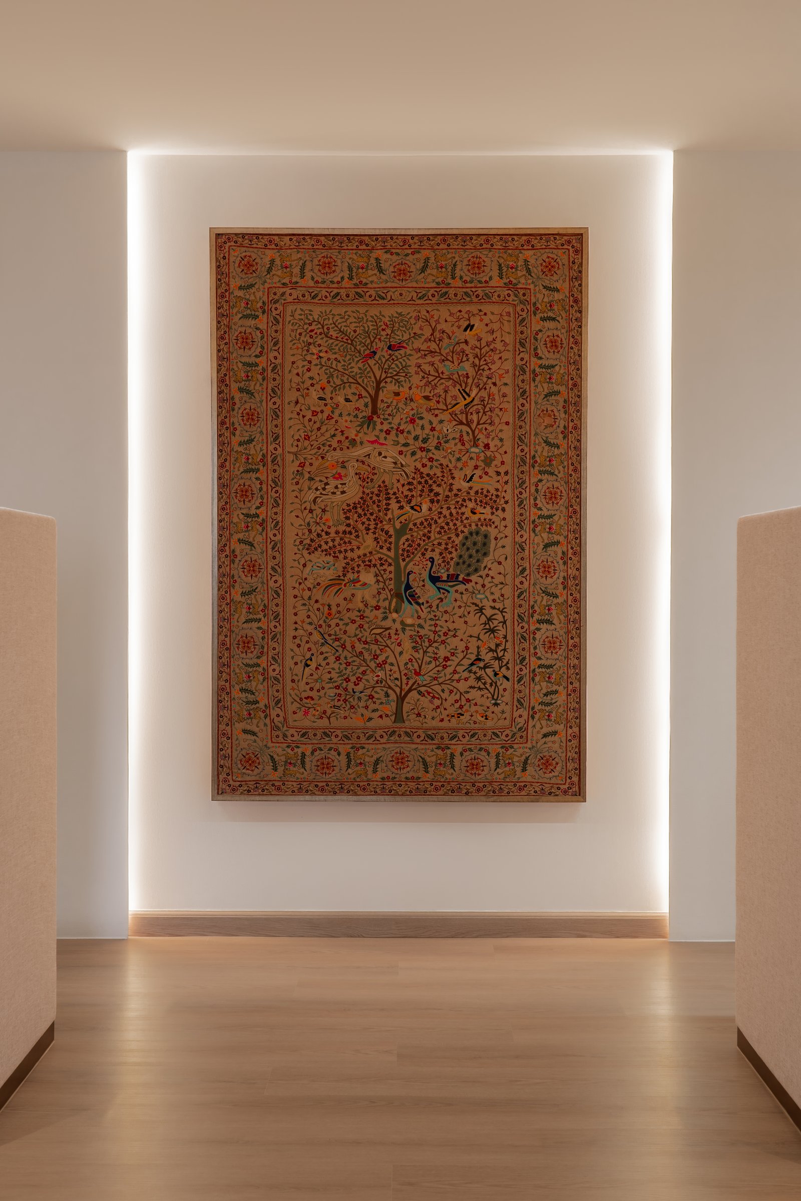 The framed textile between the reception desks, an embroidered field of birds and florals positioned as the office's cultural anchor