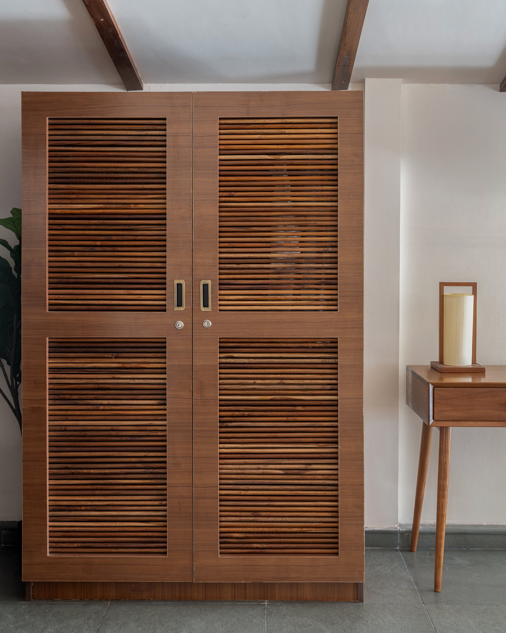The wardrobe closed, its teak louvered doors and recessed brass pulls reading as a piece of furniture in their own right