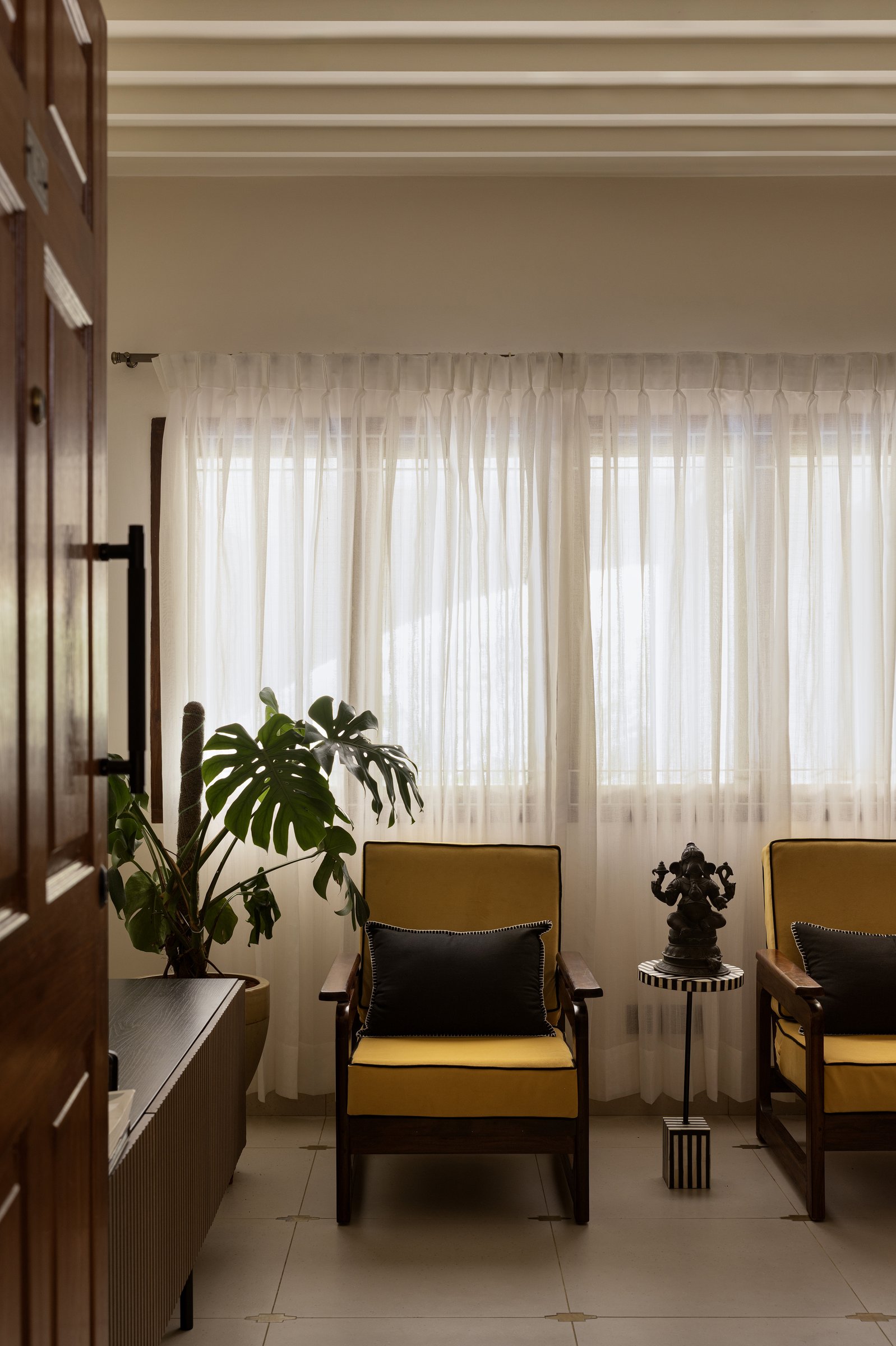 The living room's second seating group, where yellow-upholstered rosewood armchairs sit against sheer curtains that soften the daylight
