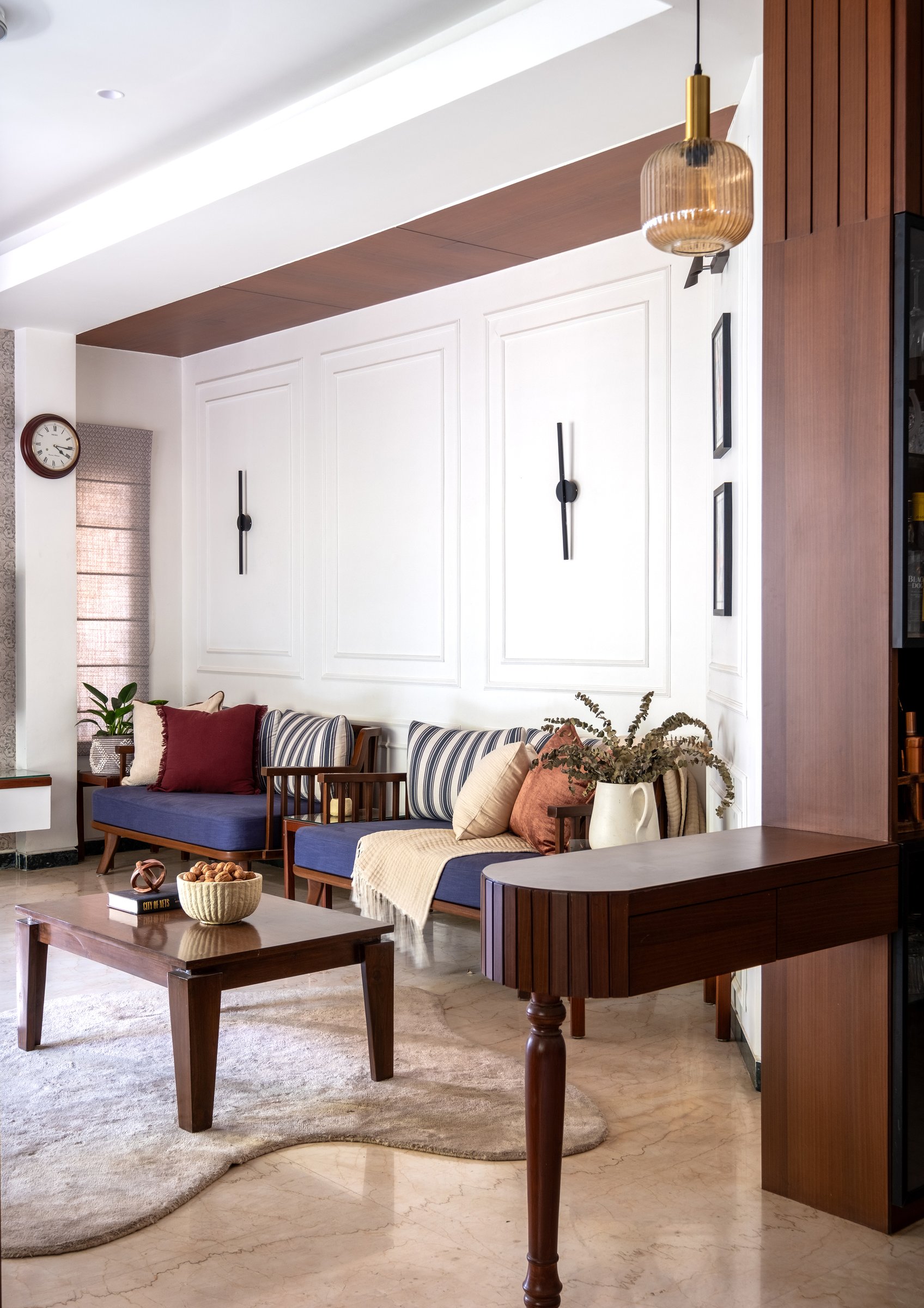 A pair of wooden daybeds with blue cushions set against a panelled wall, softened by a warm ribbed glass pendant