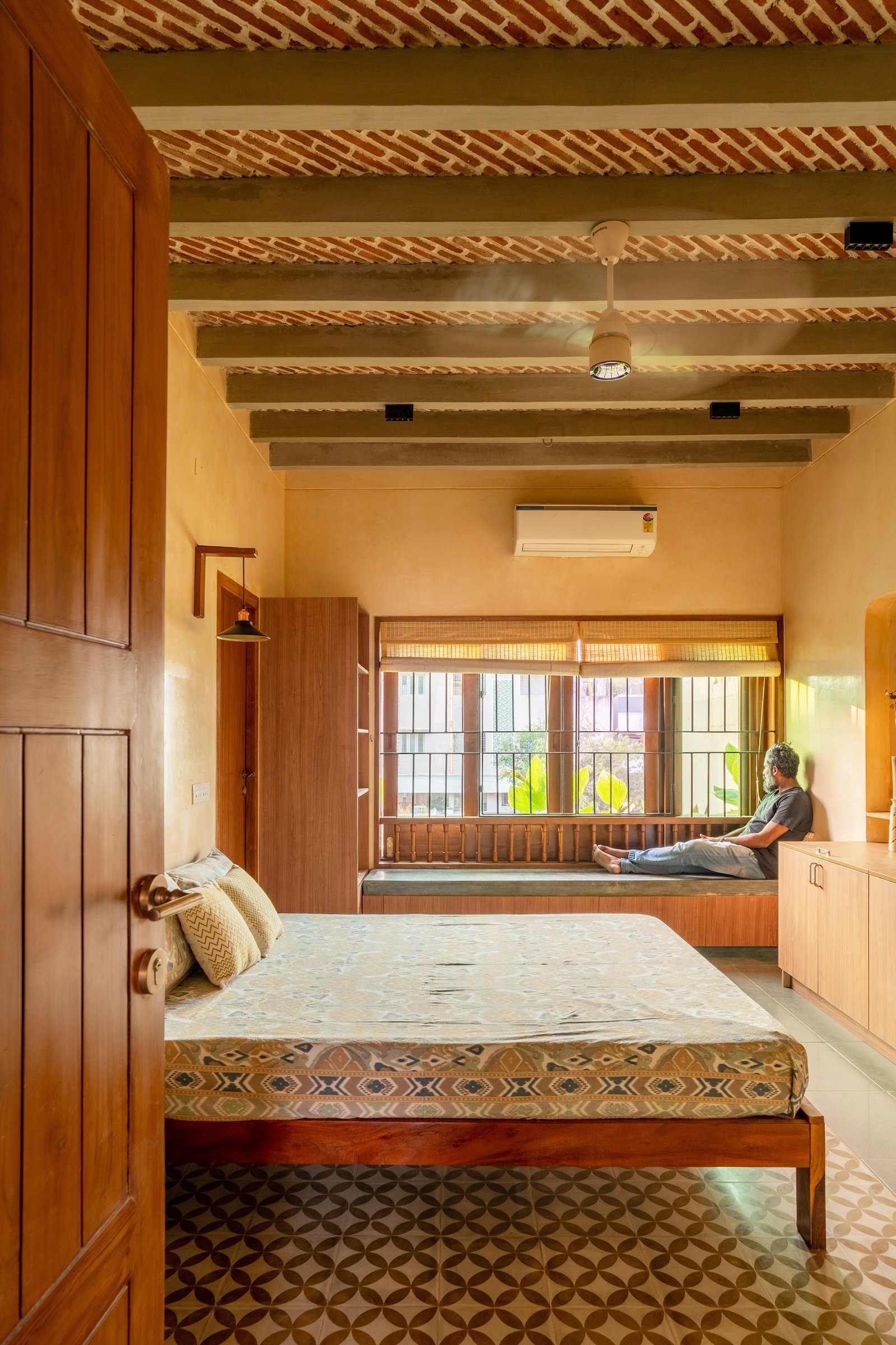 A bedroom with a low timber bed, a built-in window seat, and an exposed brick ceiling