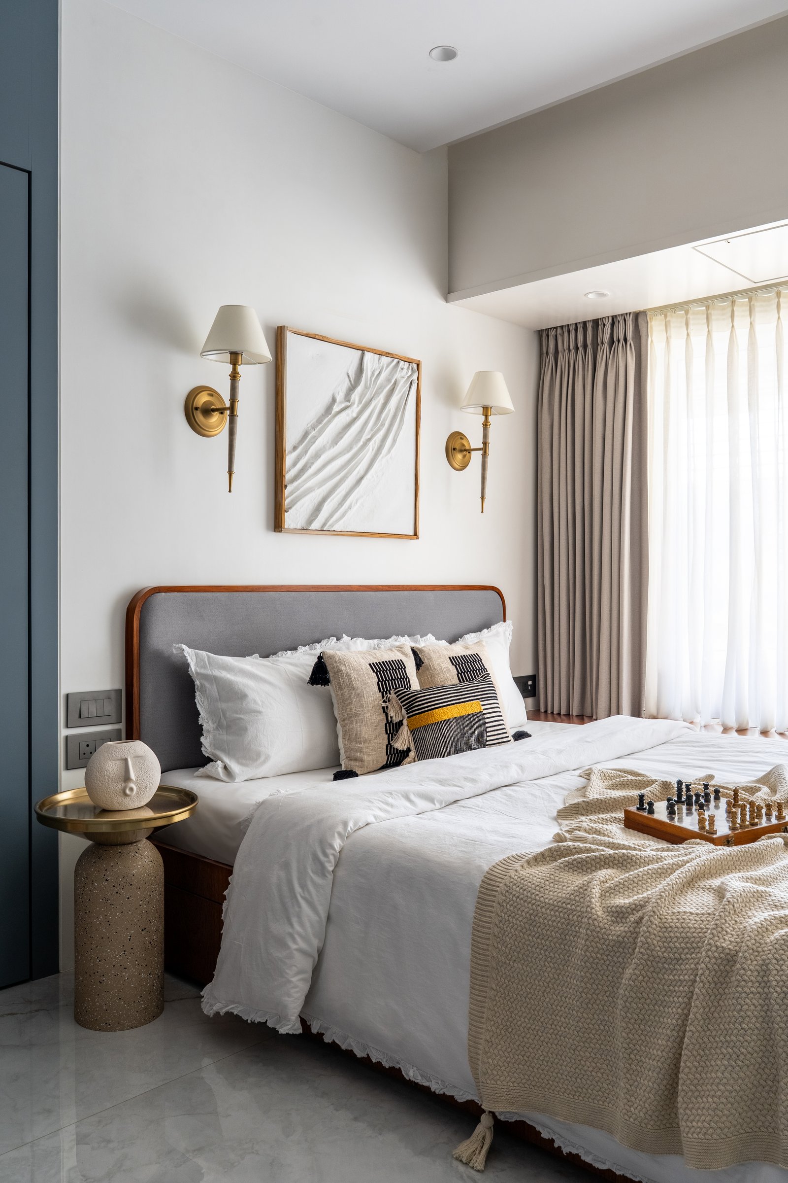 The second bedroom, where a grey upholstered headboard sits between brass sconces and a textured artwork