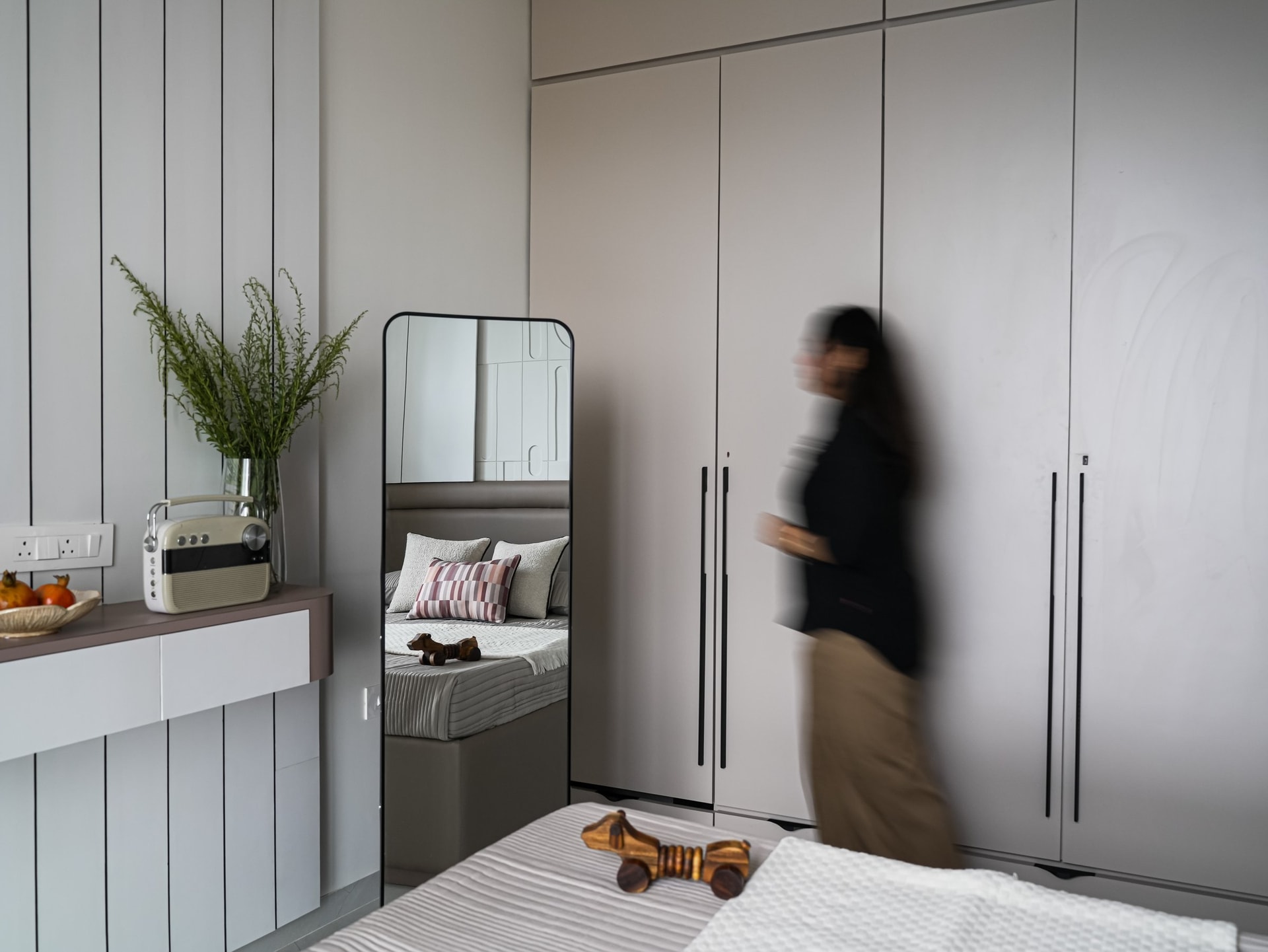 A full-height wardrobe in warm off-white recedes into the wall plane, with a leaning mirror expanding the room's perceived depth