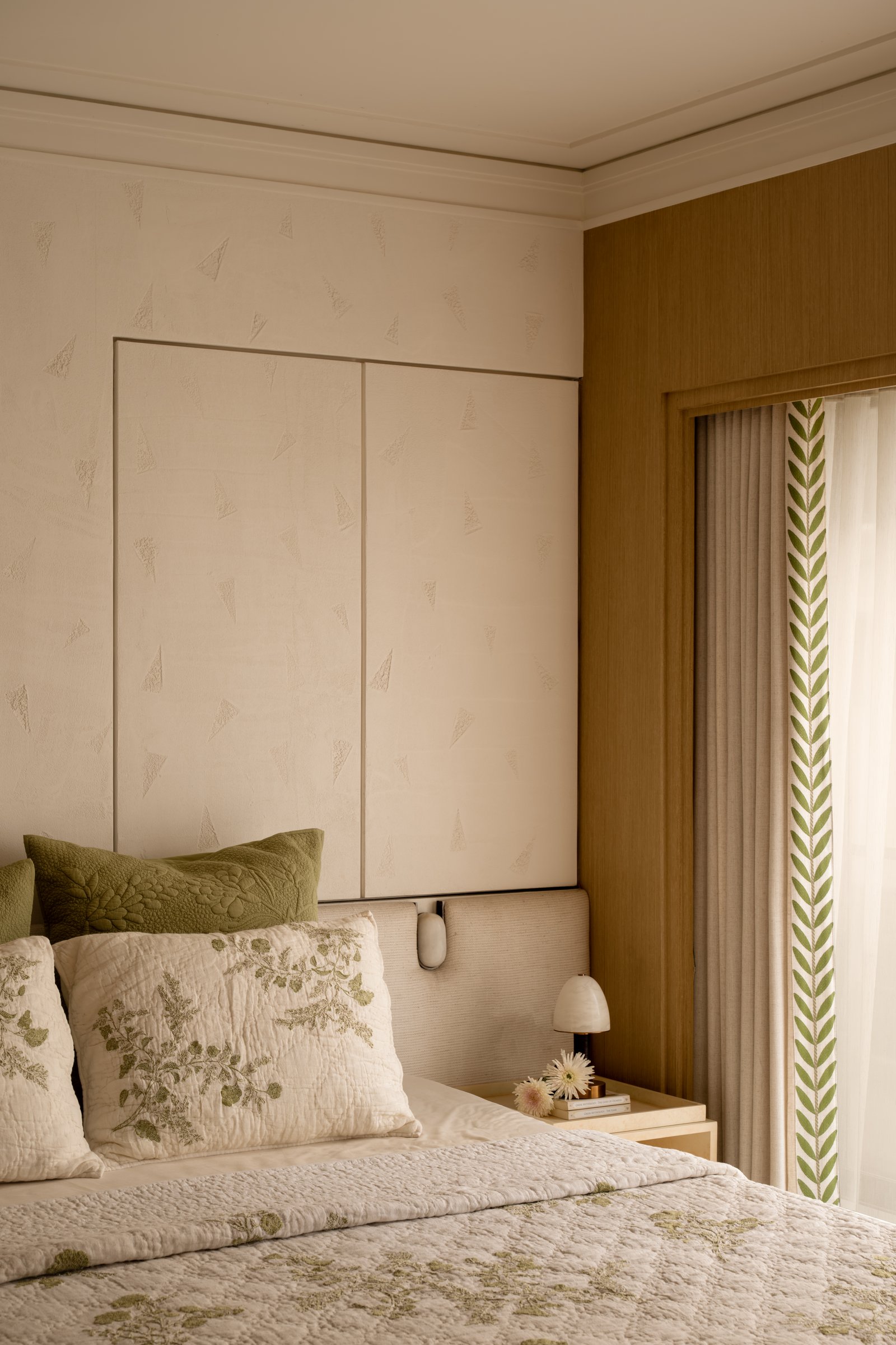 The bed wall, where a subtly patterned wallcovering and a wood-lined window reveal establish the room's tactile depth