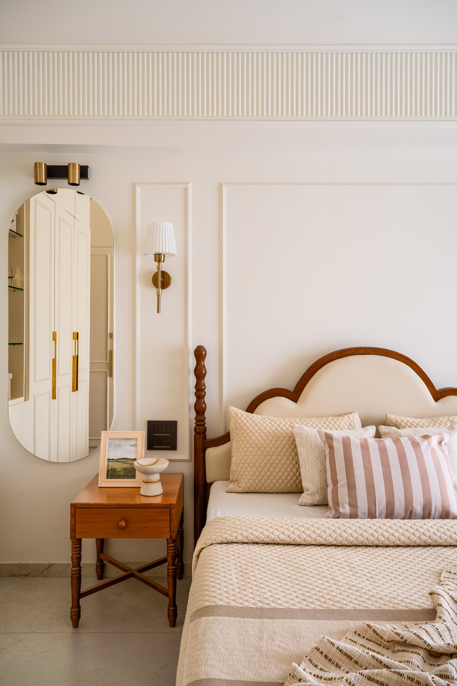 The primary bedroom, where a small hardwood side table and an oval mirror anchor a wall of raised panel moulding