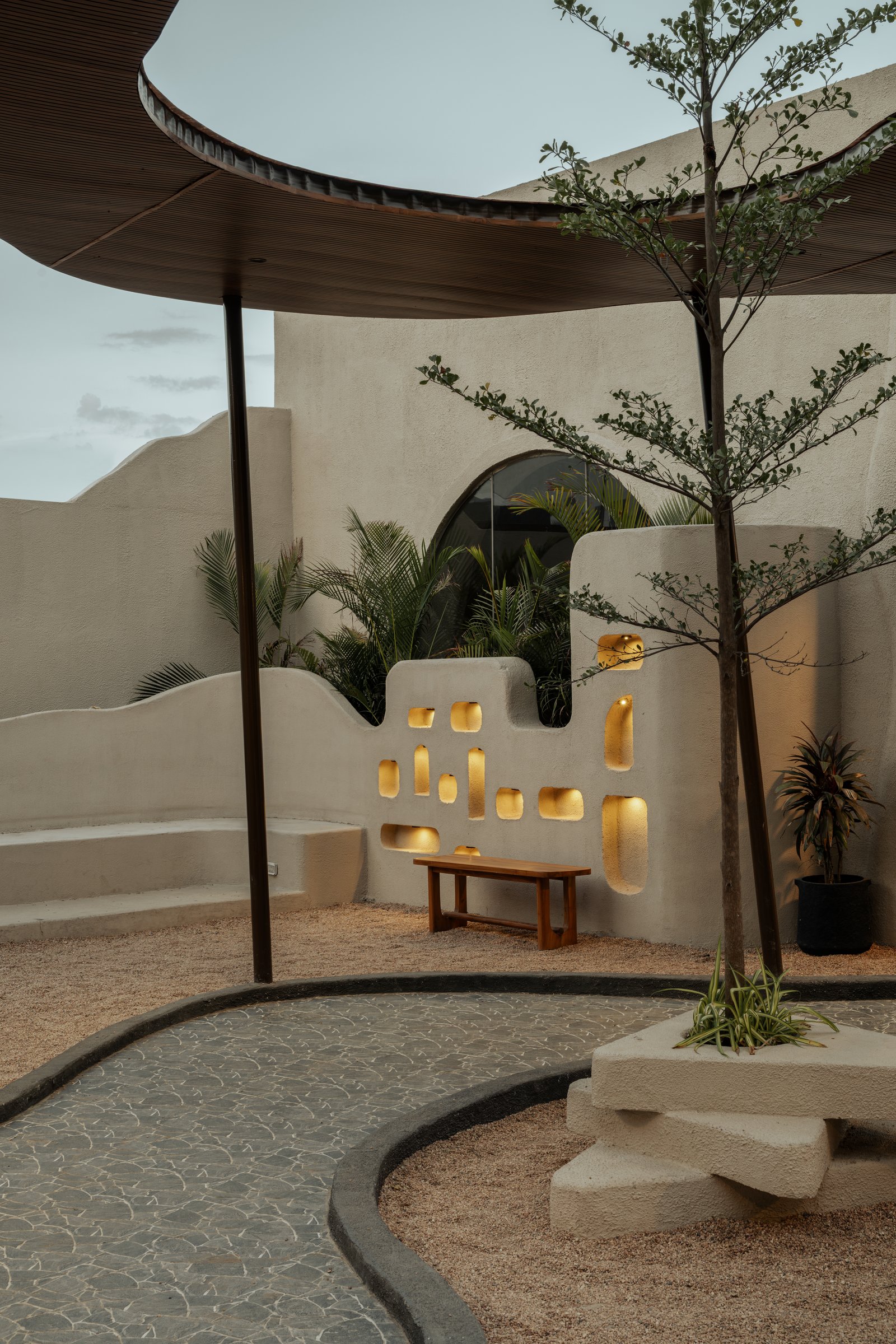 An outdoor pocket where a niched plaster wall glows amber against foliage, a low bench and stepped concrete planter completing the composition