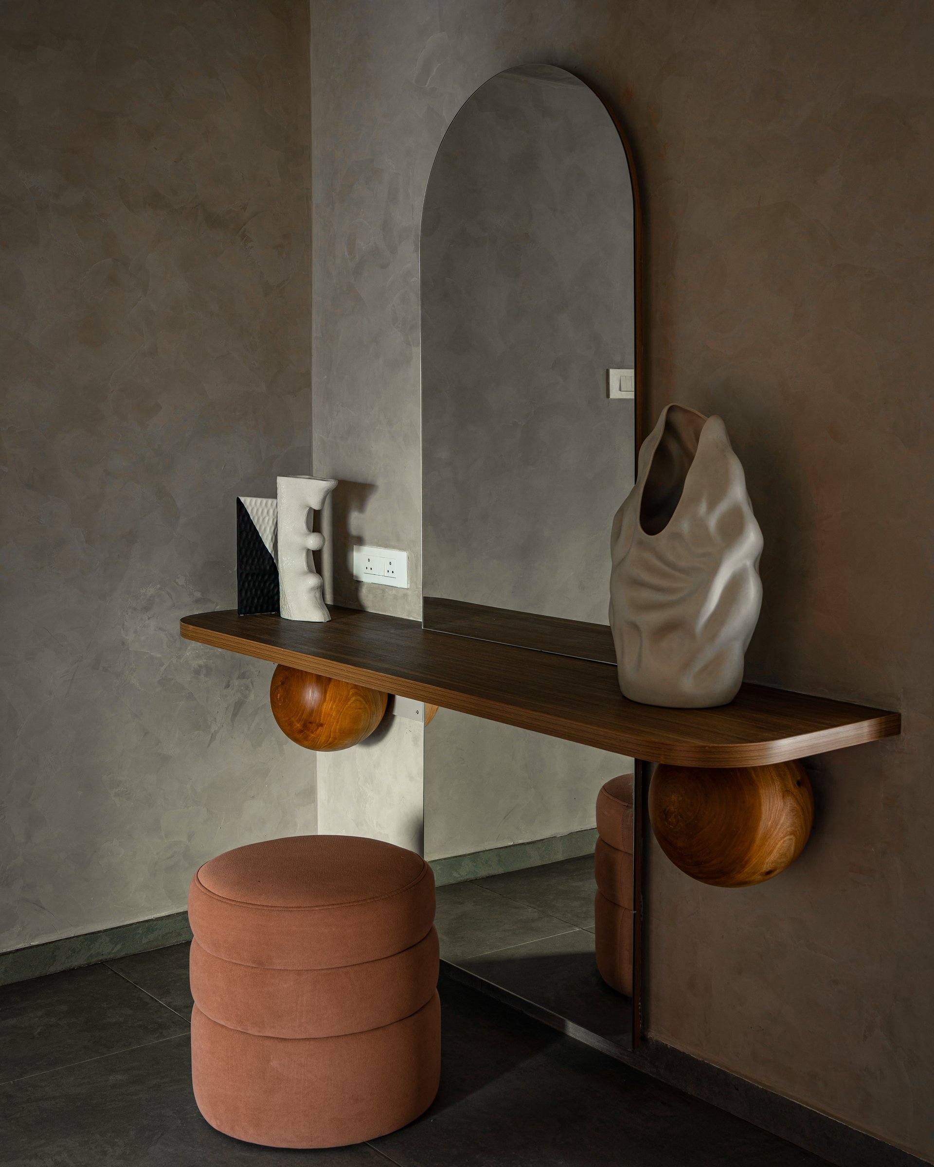The entry vignette: an arched mirror, wall-mounted wooden shelf on turned spheres, and a rounded terracotta stool set the material grammar