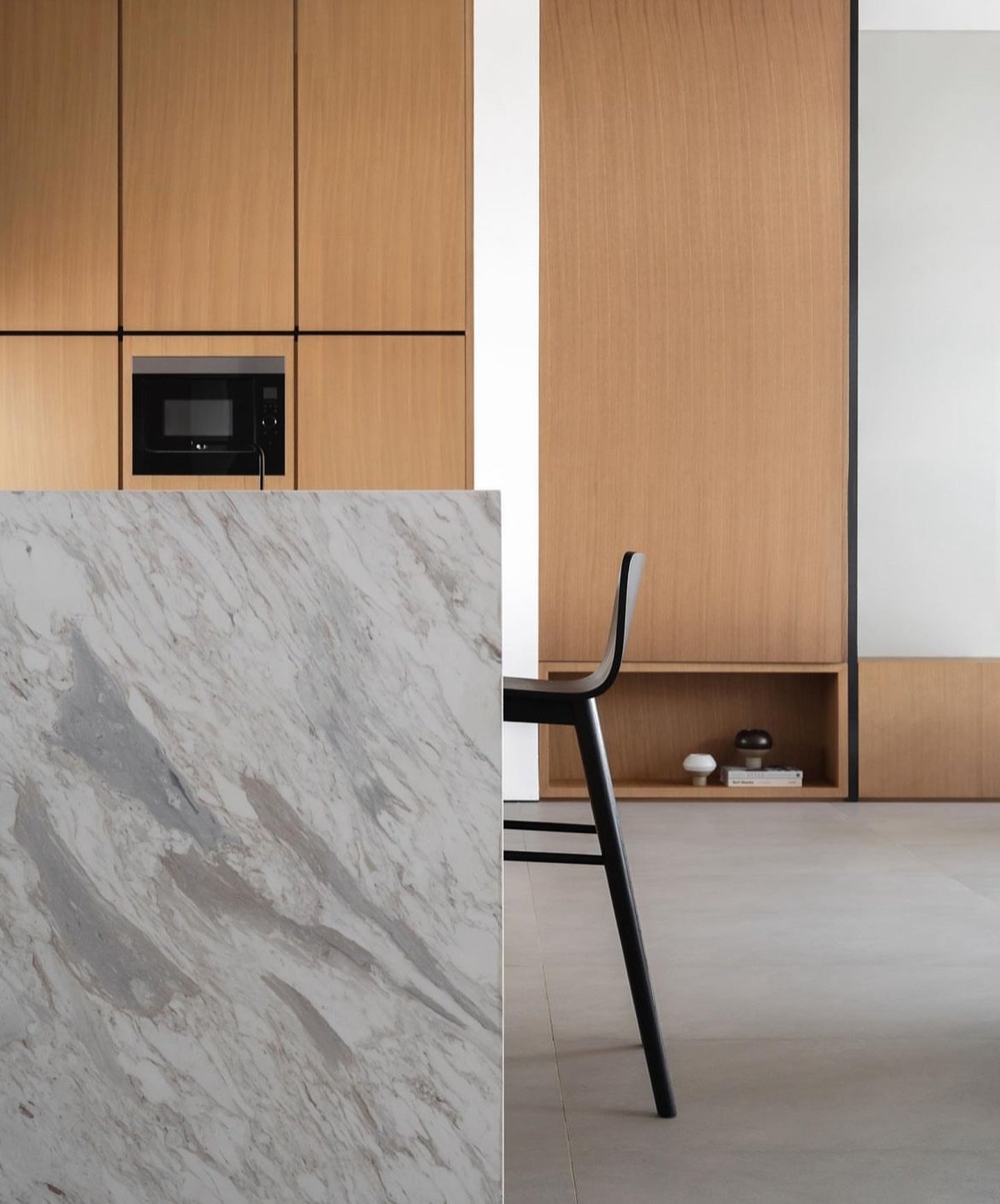A book-matched marble island meets wood-veneer cabinetry, with thin black reveals echoed in the slender legs of a single bar stool.