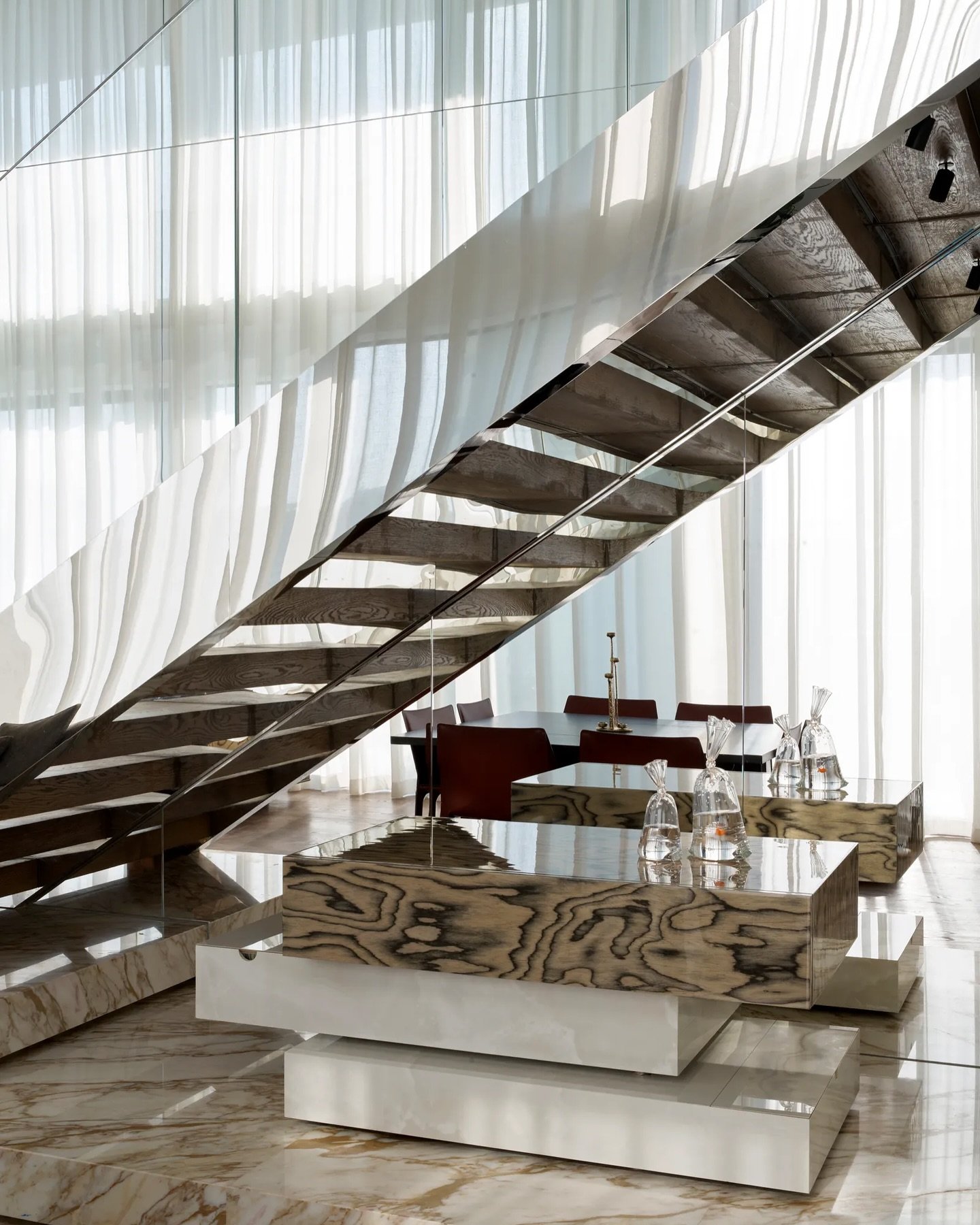 A cantilevered staircase with a polished metallic soffit reflects the dining zone, above a console of stacked wood and pale stone.