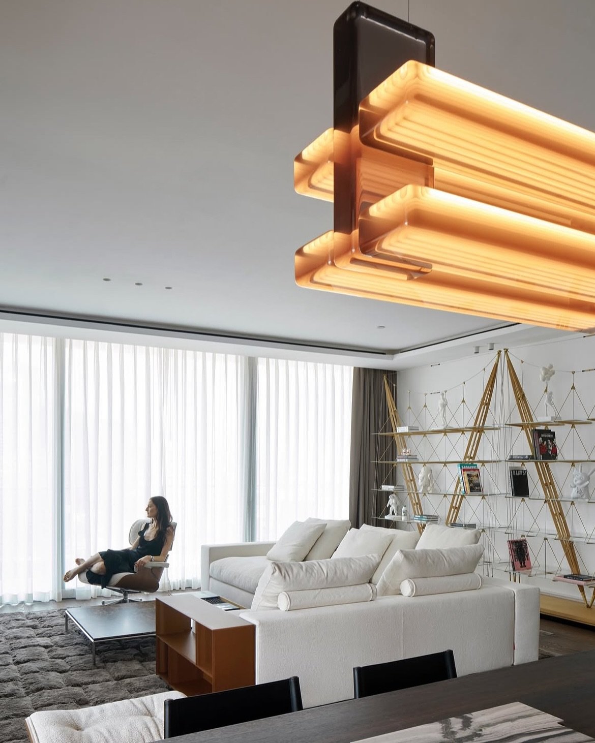 The living room gathers around a sculptural amber pendant, a brass-framed shelving unit, and a reader at the window.