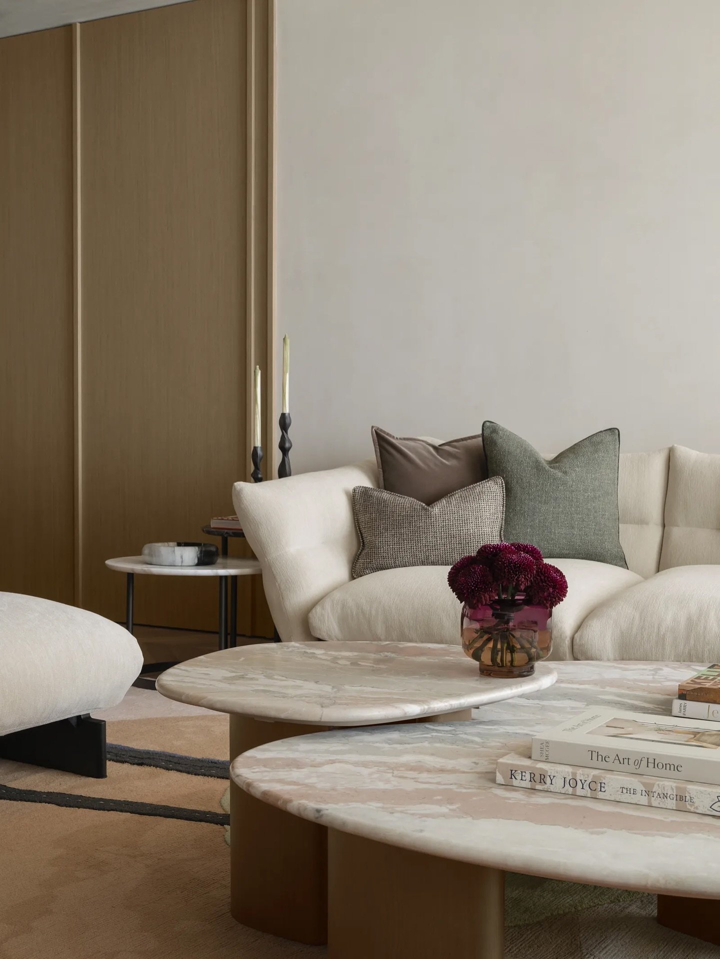 A cream sofa curves against a limewashed wall, framed by fluted wood-toned joinery and overlapping marble tables.