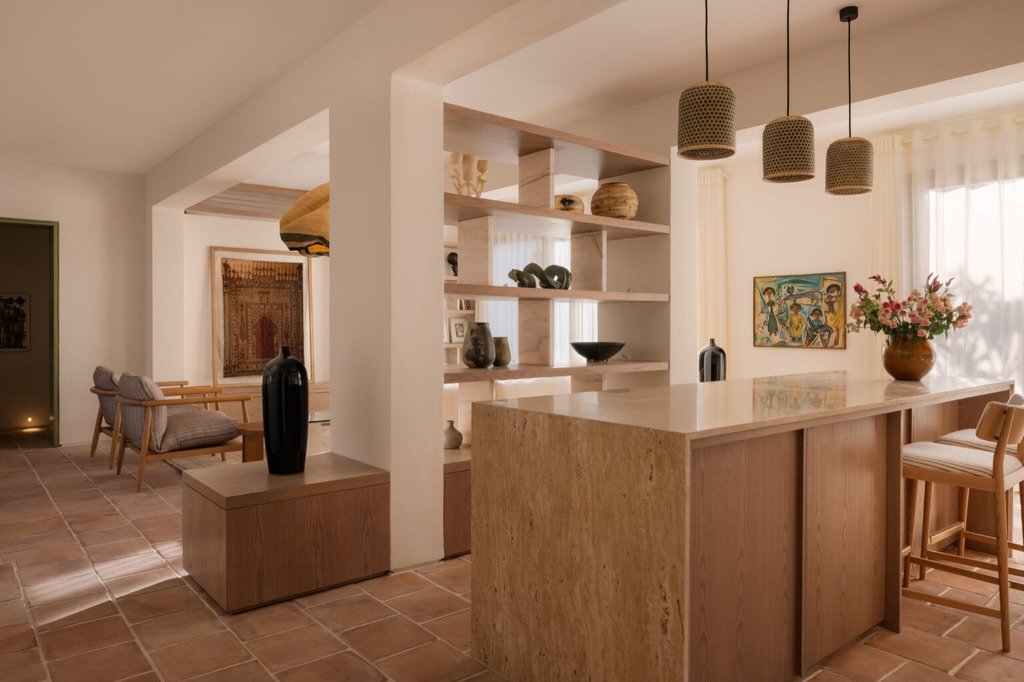 A travertine-clad island anchors the open-plan kitchen, its cane pendants and shelved ceramics quietly articulating the space.
