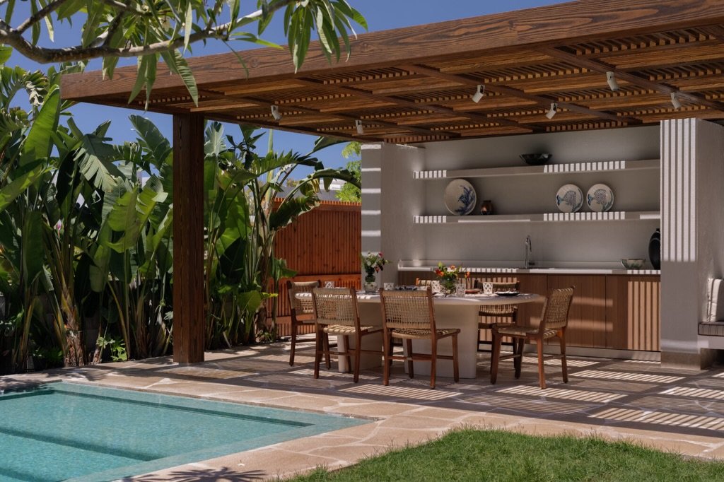A slatted pergola filters midday sun over the poolside dining area, tying stone, rattan, and foliage into a single rhythm.