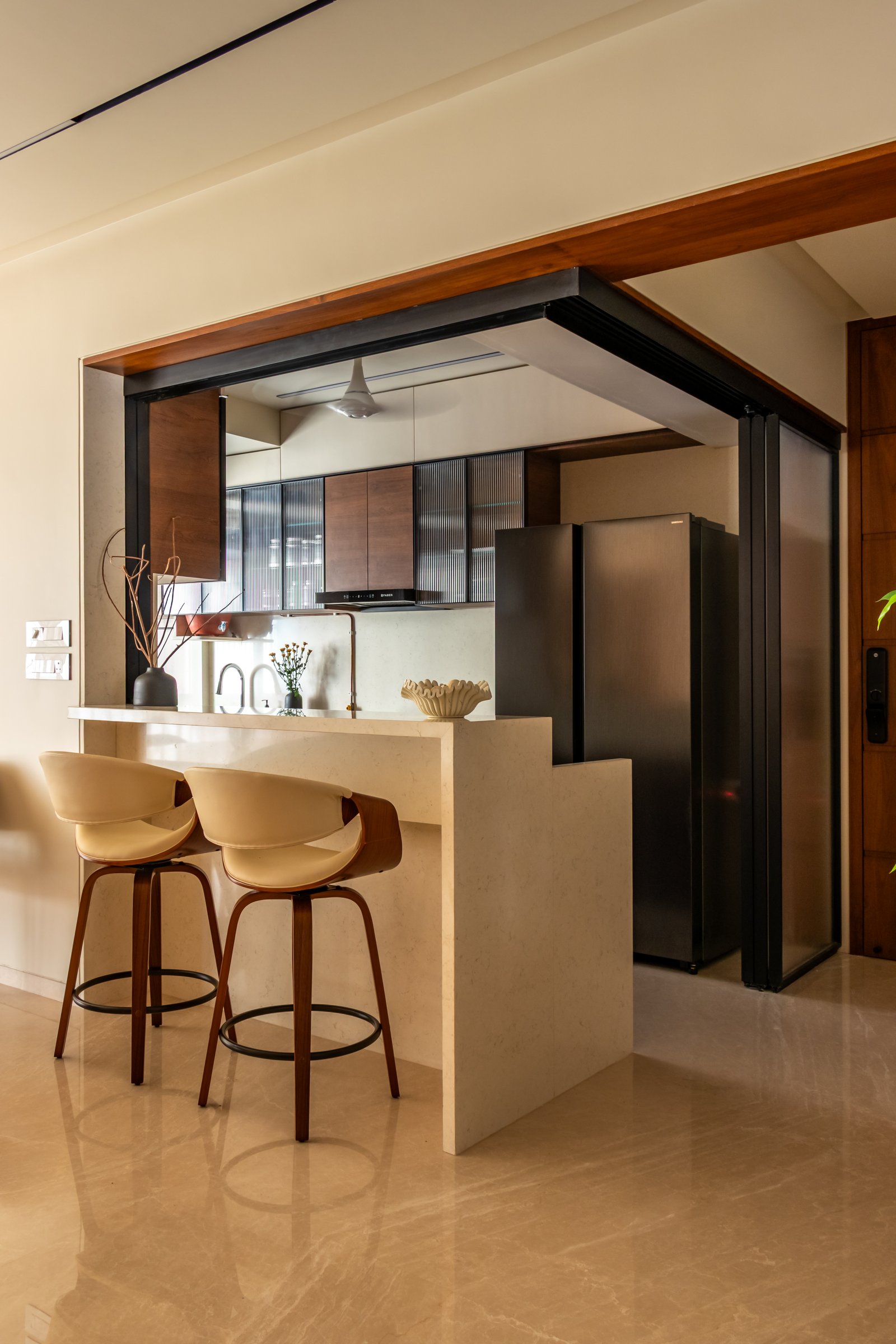 The kitchen threshold, framed by a metal-and-hardwood portal, with reeded-glass uppers and warm veneered shutters visible beyond the breakfast counter