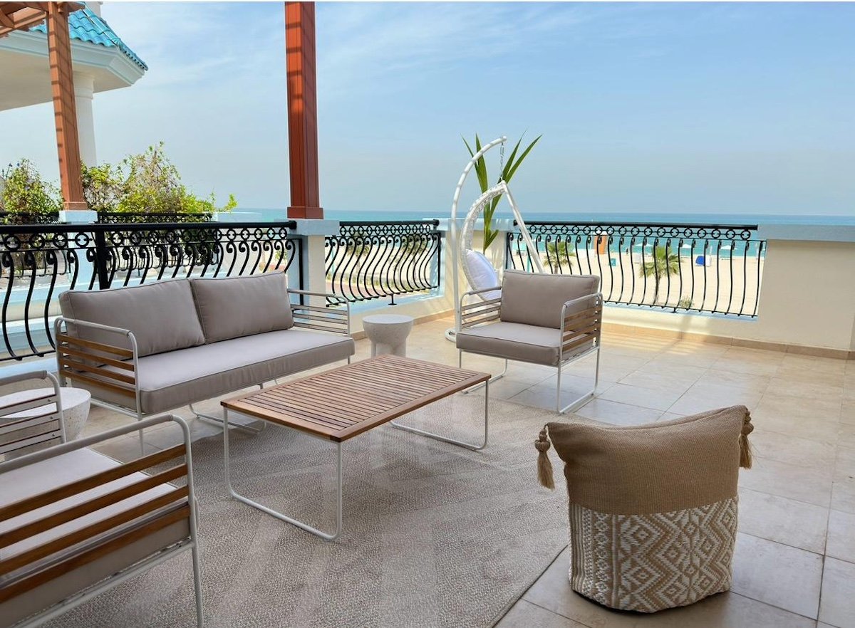 The terrace, where slatted teak, white steel, and a woven basket in ivory and jute extend the villa's coastal palette into the open air