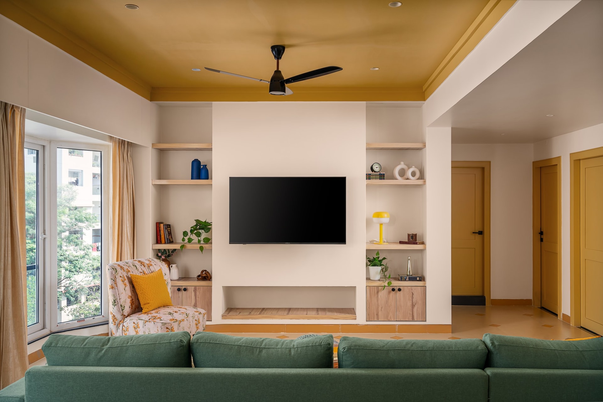 The media wall composed as architecture, with open wooden shelves flanking the screen and an upholstered armchair by the window