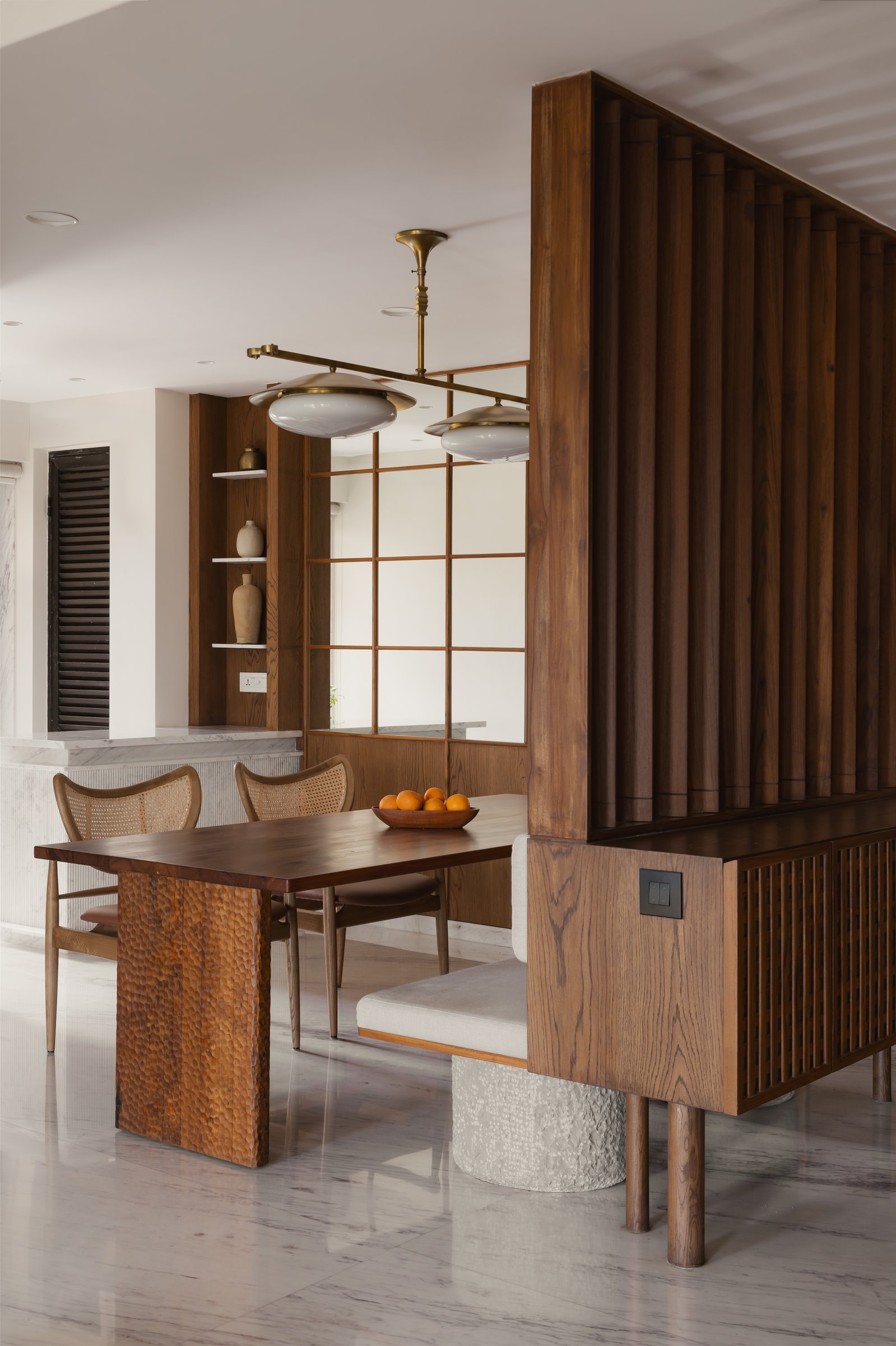 The handcrafted teak dining table on its hammered pedestal, paired with a sculptural brass-and-opal light fixture overhead