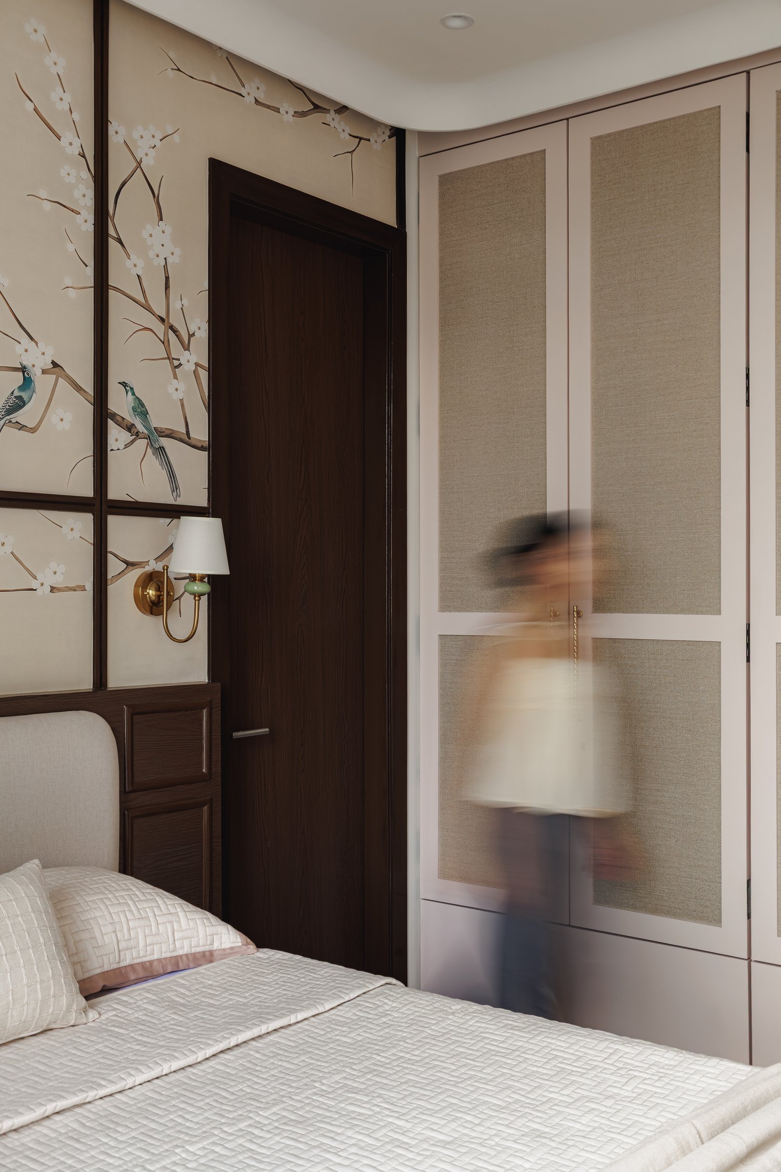A view across the primary bedroom, where linen-finish wardrobe panels in dusty blush meet the darker panelled wall