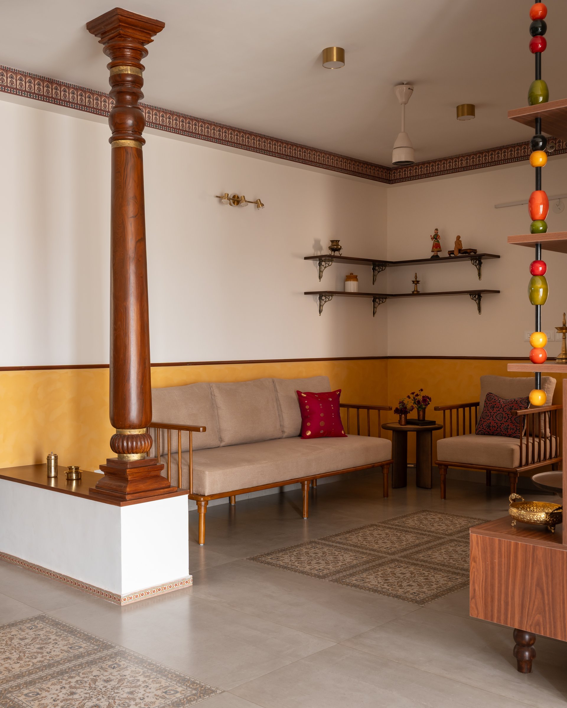 A low wooden-framed sofa and armchair rest against the mustard wall, with brass vessels displayed on scrolled corner shelves