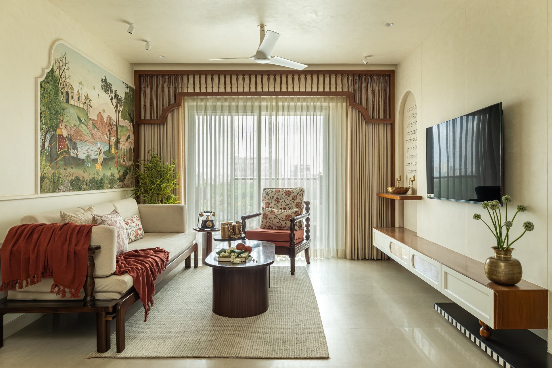 The living room, where a Mughal miniature-inspired mural anchors the seating composition and a carved jharokha-style valance frames the balcony