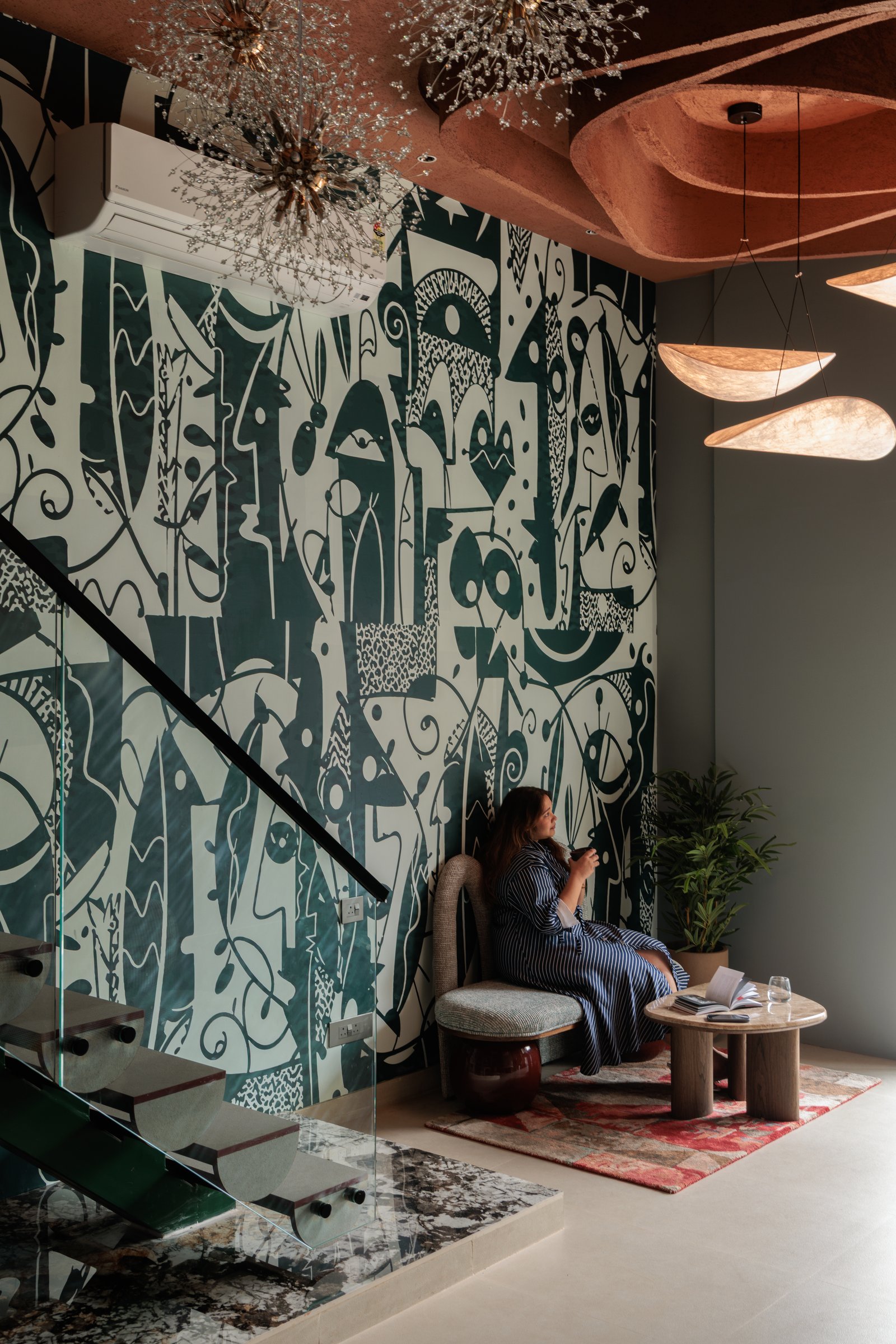 The double-height mural rising alongside the stair, with sculptural pendants suspended against a terracotta-toned ceiling recess