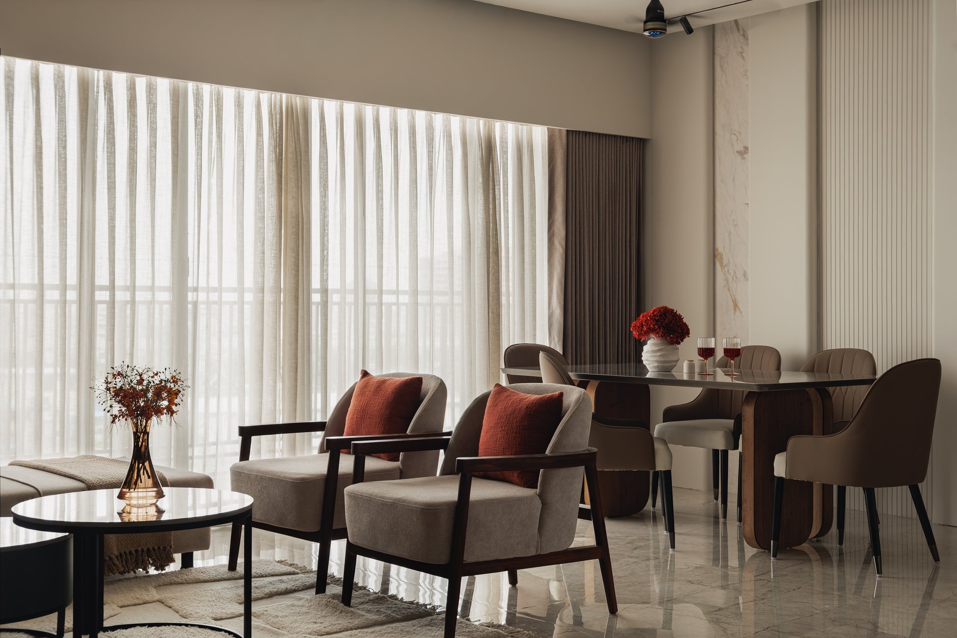 A wider view connecting the dining area with the adjacent lounge seating, where timber-framed armchairs sit beside sheer-curtained windows